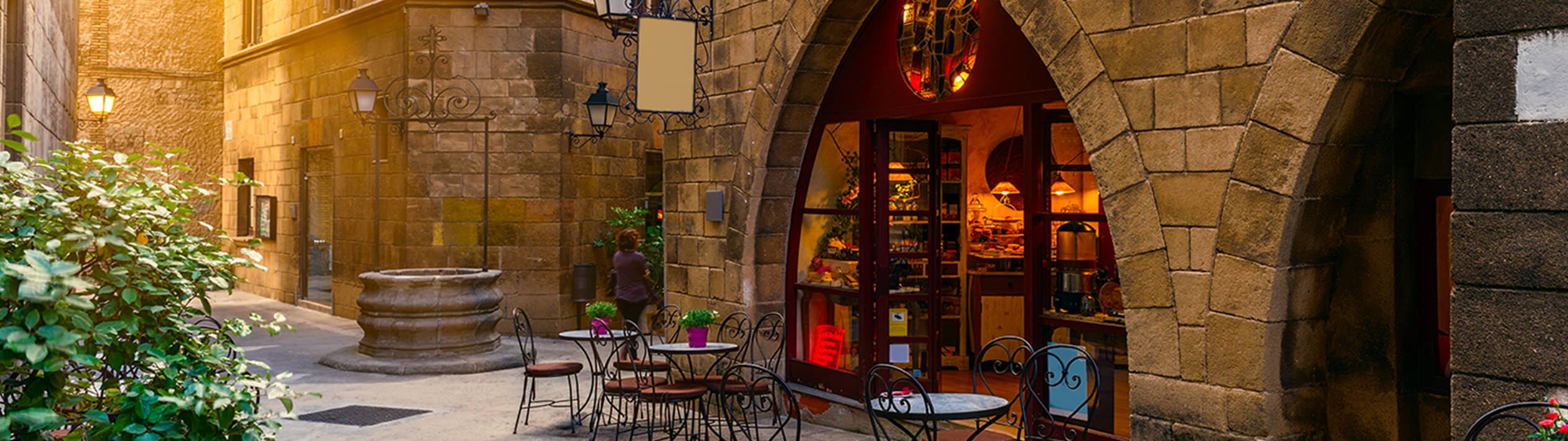 In Barcelona, a charming courtyard with tables and chairs sits in front of a verdant, flower-adorned building. A figure stands amidst twinkling lights and boards, while inside, a cupboard holds various objects.