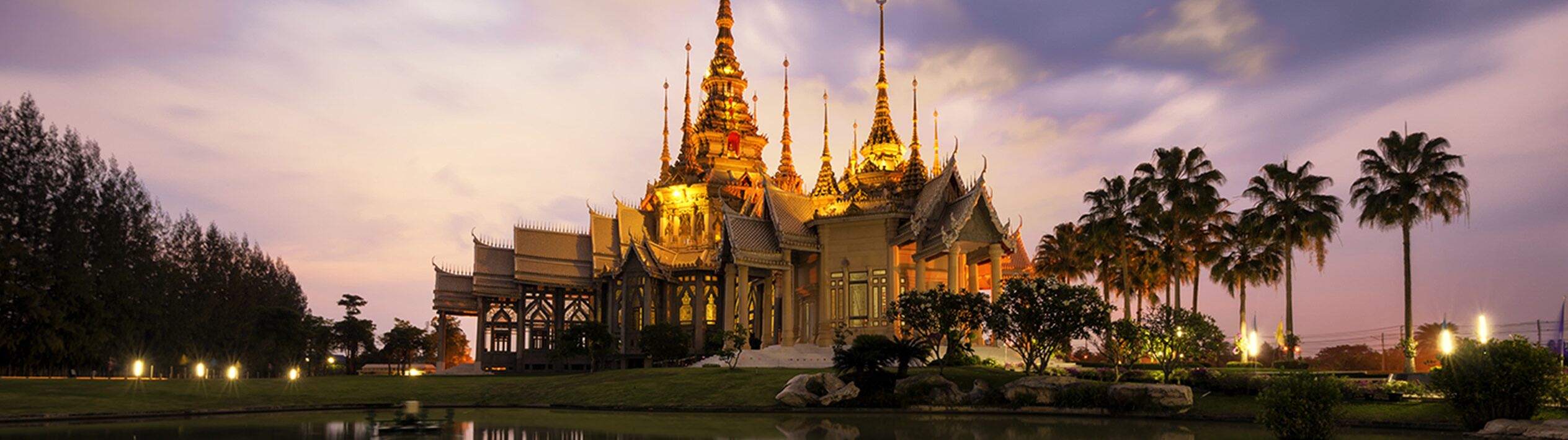 In Bangkok, Wat Phra That Doi Suthep shines brightly in the night. Lush greenery and a calm lake surround it, while clouds drift over the illuminated temple, creating a serene and majestic view.