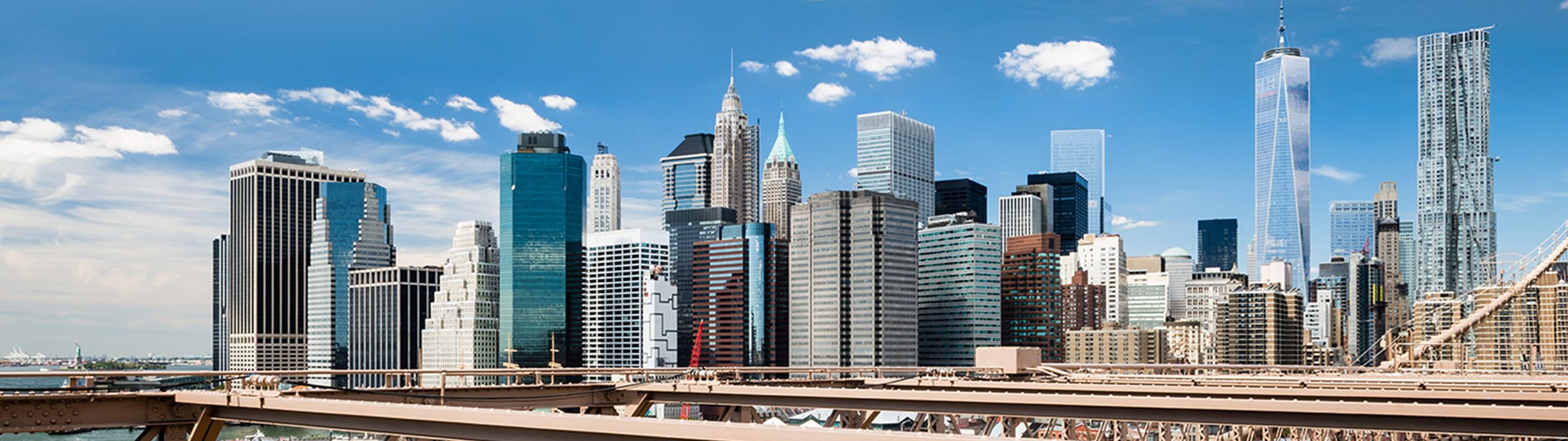 In New York, from the Brooklyn Bridge, the Manhattan skyline rises against a cloudy sky. Below, boats drift across the water, capturing the city's vibrant pulse and iconic beauty in a single scene.