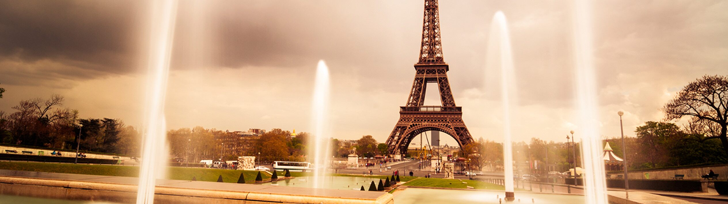 In Paris, the Eiffel Tower stands tall amid a park, surrounded by a fountain, lush green grass, trees, and light poles. Nearby, people and vehicles move under a sky dotted with fluffy white clouds.