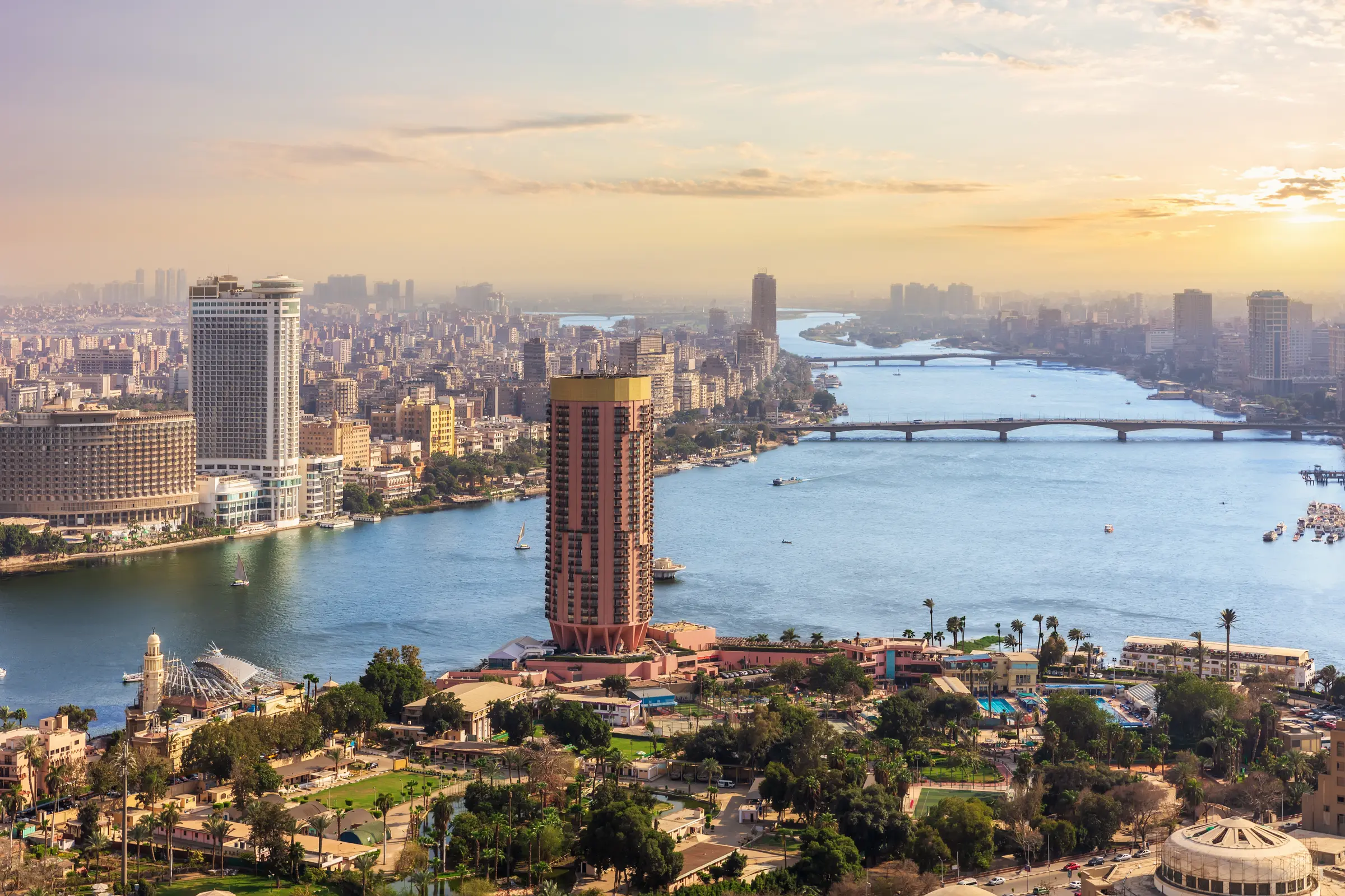 In Cairo, the Nile River glistens at sunset. Buildings, trees, and bustling vehicles line the road. A bridge spans the river under a cloud-filled sky, with the sun setting beautifully beyond.