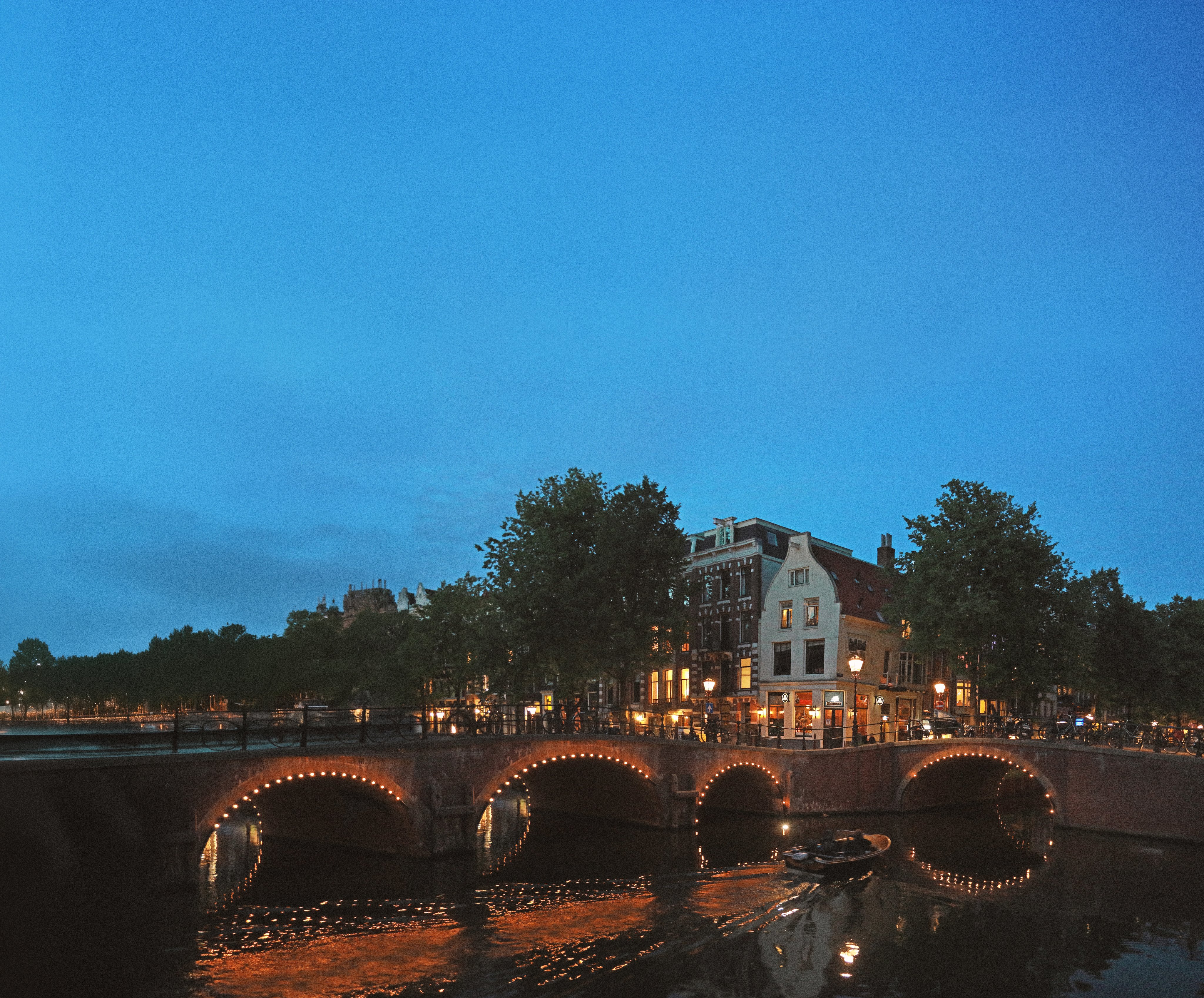 In Amsterdam, a bridge glows over a canal at night. A boat glides beneath, embraced by lit trees and buildings. The backdrop of a starry sky completes this serene, picturesque scene.