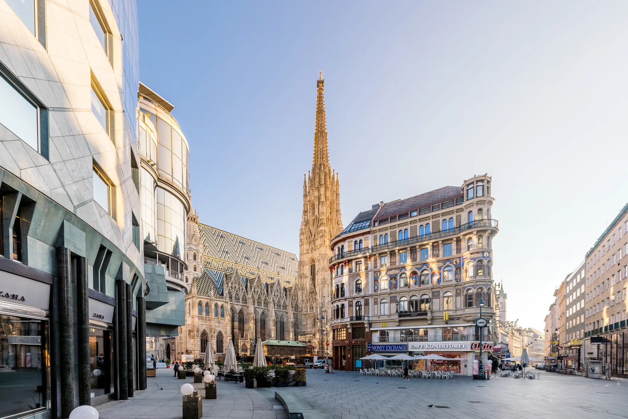 In Vienna, a bustling street scene unfolds with a towering building in the backdrop. Street lights illuminate tables and chairs scattered under umbrellas, with plants adding charm beneath the open sky.