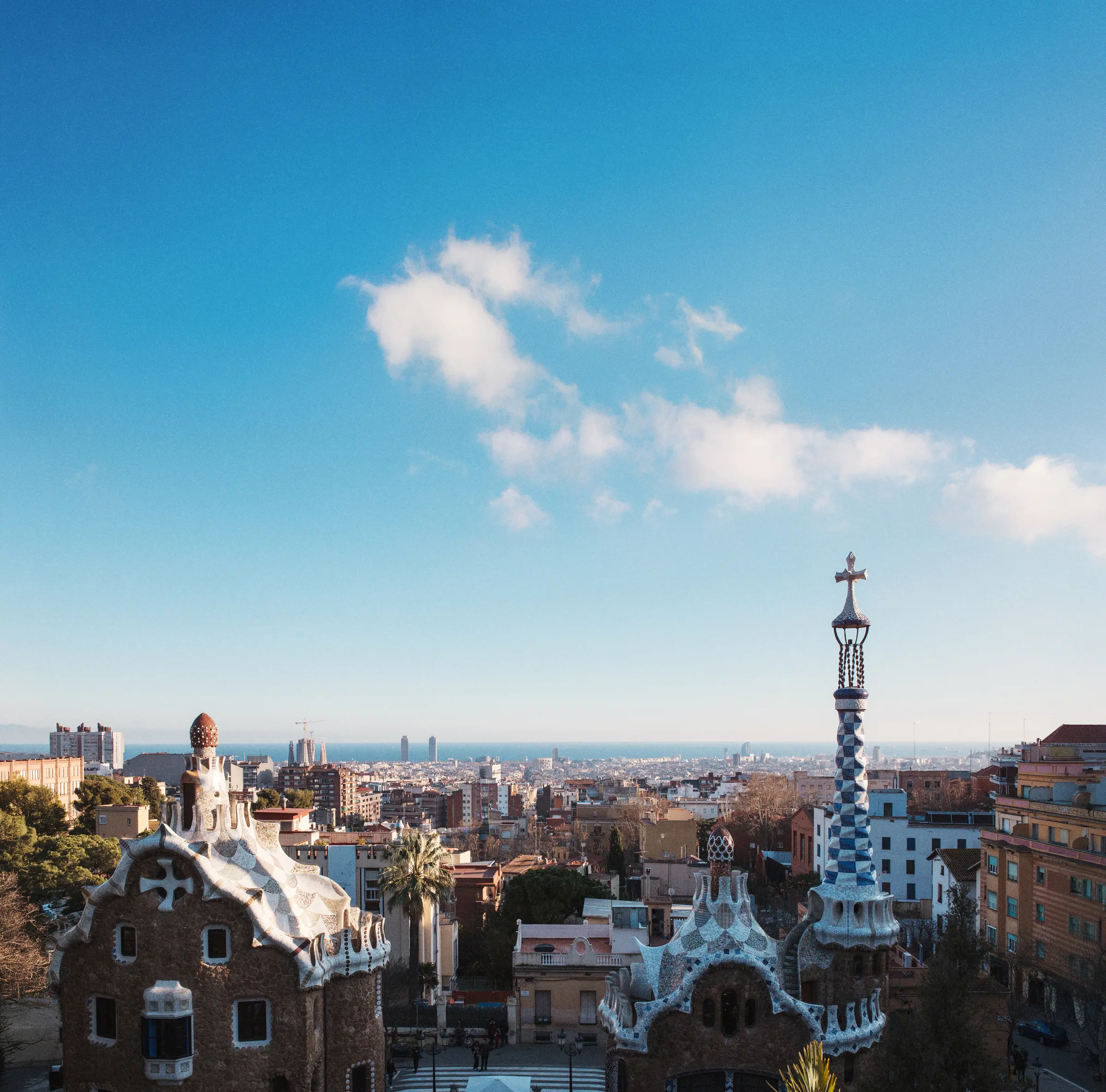 From a hilltop in Barcelona, the view unfolds: buildings and trees stretch across the landscape. Below, a few people walk along the road. Above, the sky is majestically filled with clouds.