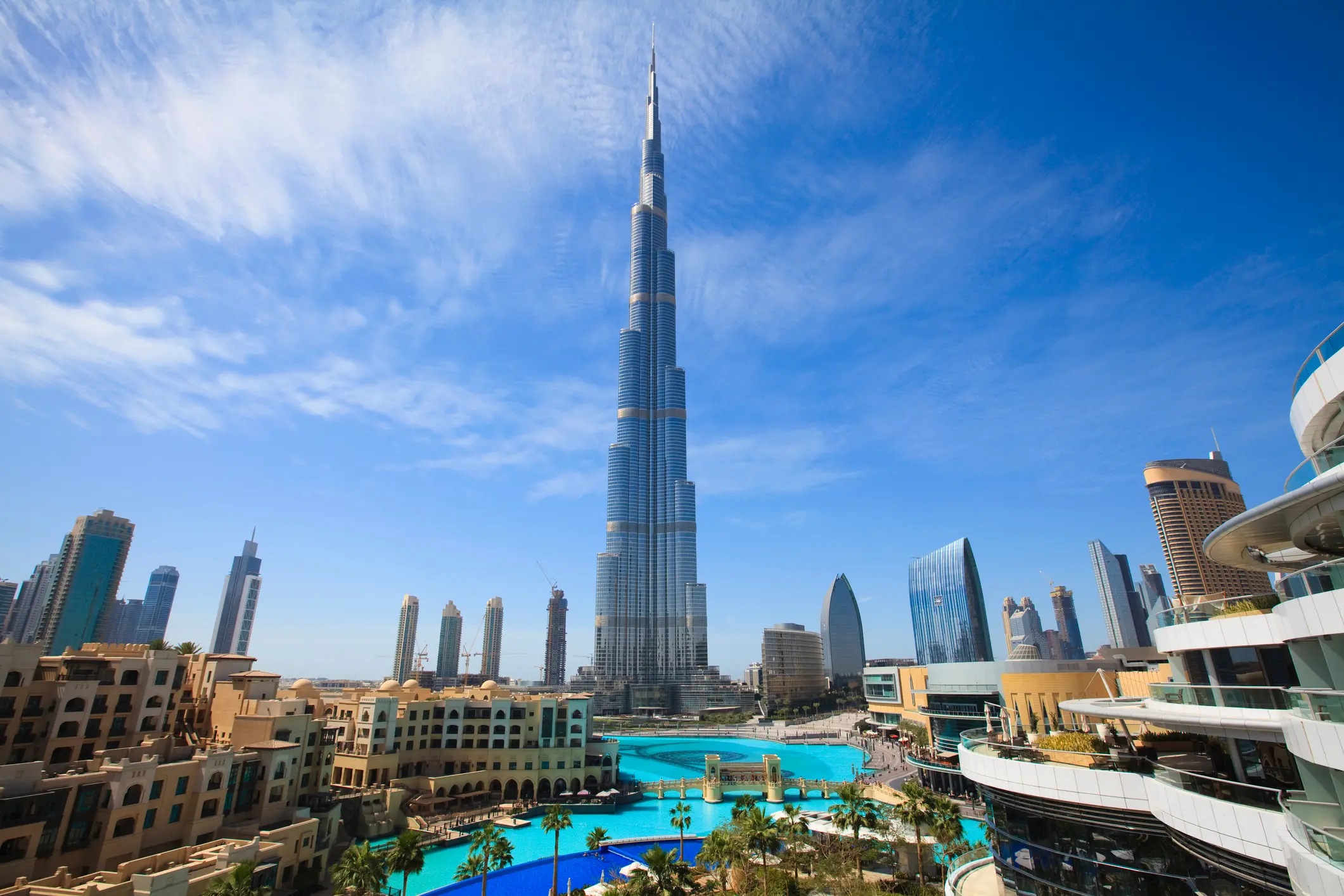 In Dubai, the Burj Khalifa pierces the cloudy sky, surrounded by trees, buildings, and a serene swimming pool, crafting a stunning urban oasis against the dramatic celestial backdrop.
