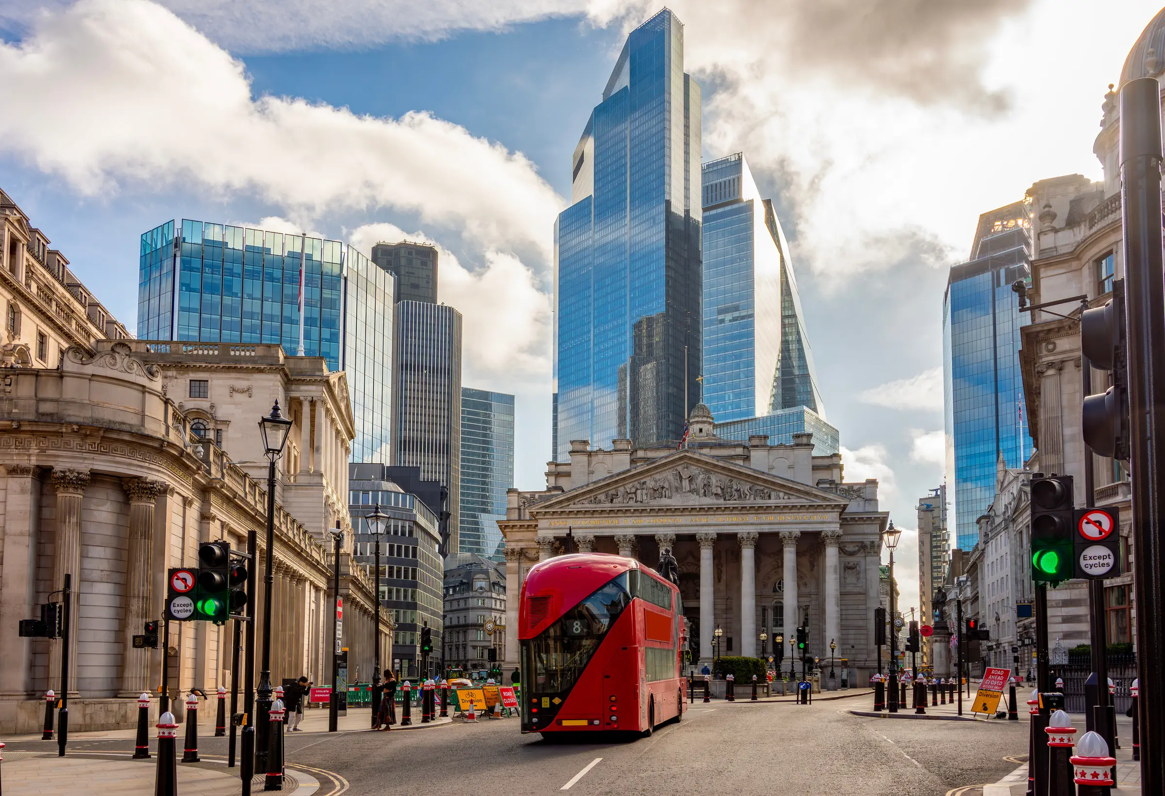 A red double-decker bus glides through a London street, flanked by towering buildings. Poles, signals, and signs line the path, while pedestrians stroll under a cloud-filled sky.
