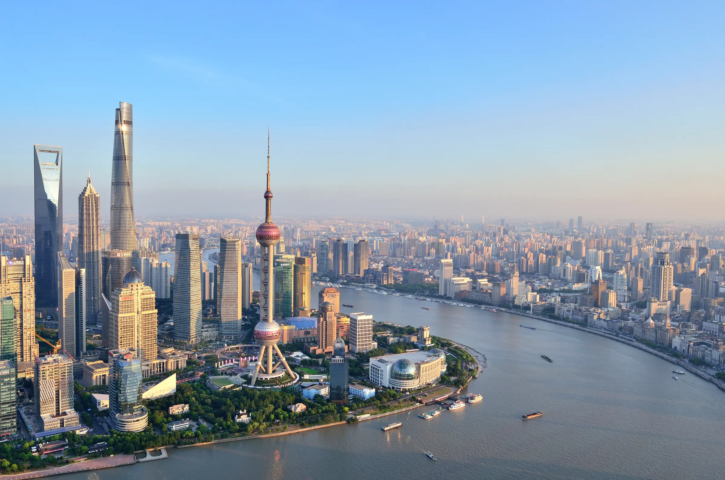 Shanghai's skyline dazzles with towering buildings, lush greenery, and shimmering waters. Boats glide through, while the sky forms a picturesque backdrop to this vibrant Chinese metropolis.