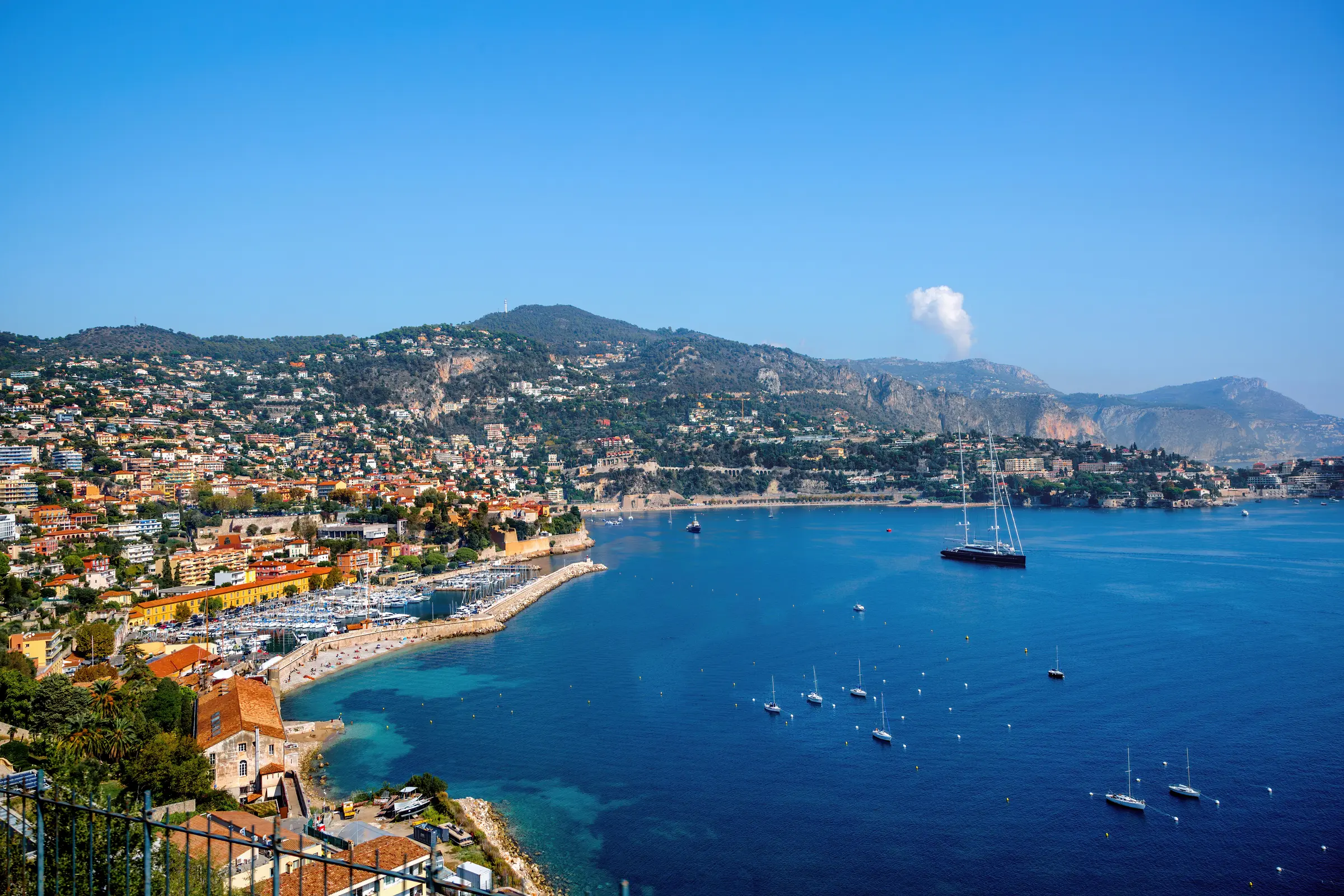In Nice, the harbor is alive with boats, embraced by buildings and trees, with majestic mountains as a backdrop. The scene unfolds under a brilliant blue sky, offering a perfect coastal vista.