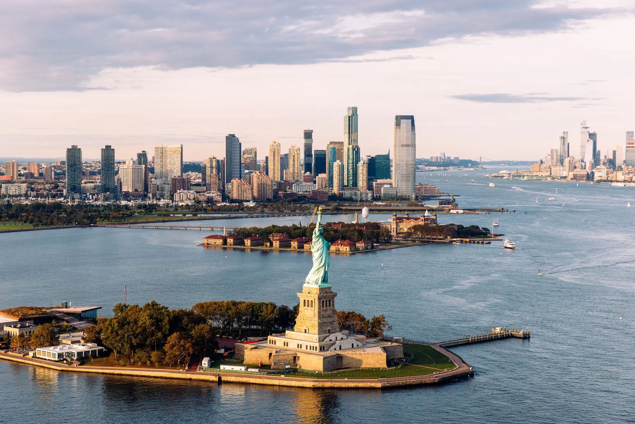 In New York, the Statue of Liberty stands majestically amid water. Boats navigate around, with trees and city buildings nearby. The sky, adorned with clouds, enhances the iconic scene's beauty.