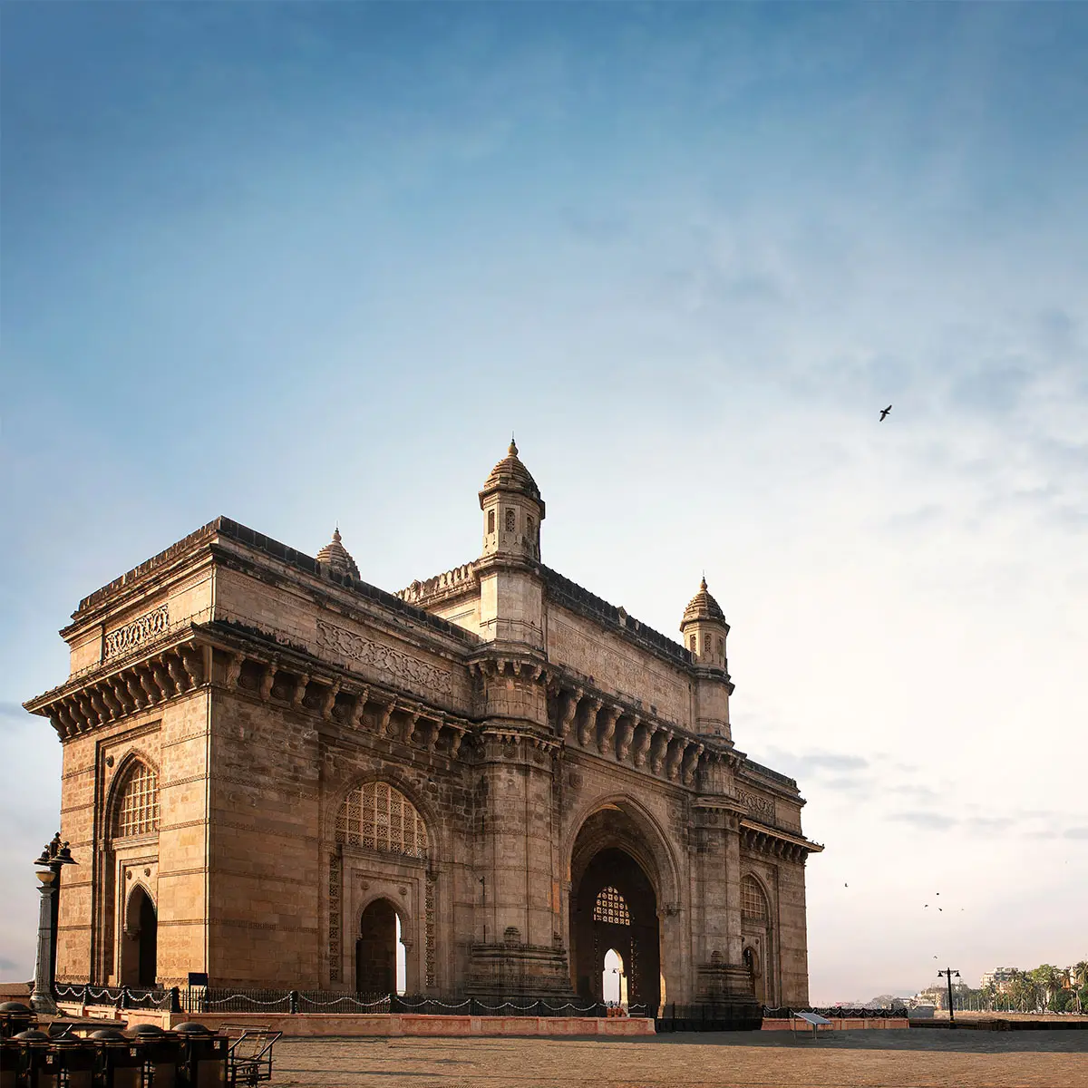 The image depicts the Gateway of India, located in Mumbai, a historic monument fenced off, with onlookers nearby. The scene includes ground objects, trees, and buildings under a clear blue sky.