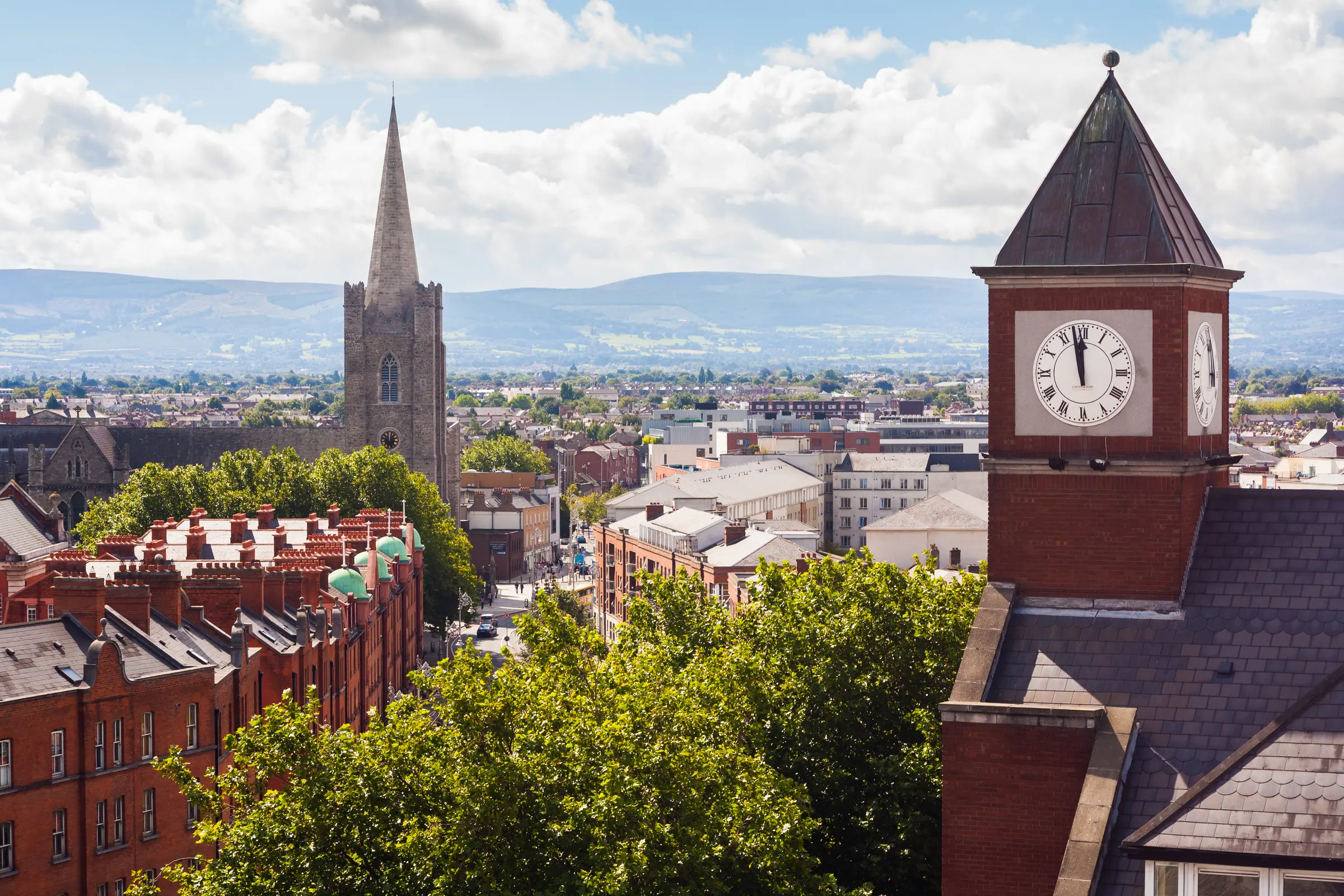 In Dublin, a cityscape features a clock tower amid bustling streets, framed by buildings, trees, and vehicles. Poles dot the area, while distant hills rise under a cloud-strewn sky.