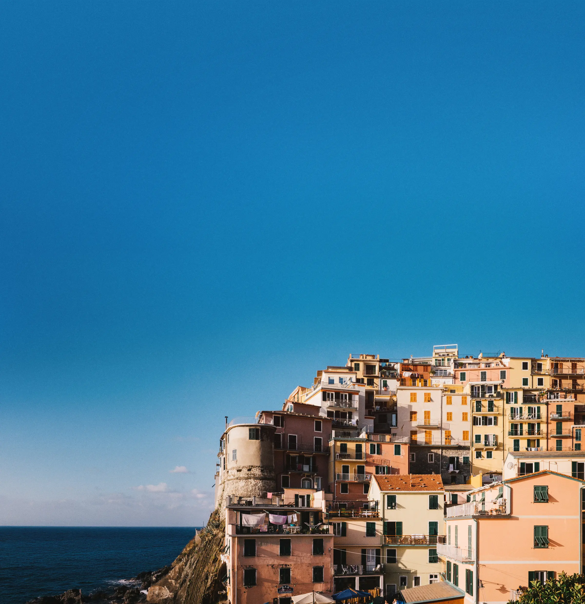 In Liguria, Italy, Manarola's vibrant buildings sit on cliffs above the ocean. The colorful mix of white, cream, and brown structures contrasts beautifully with the deep blue-green water and brilliant sky.