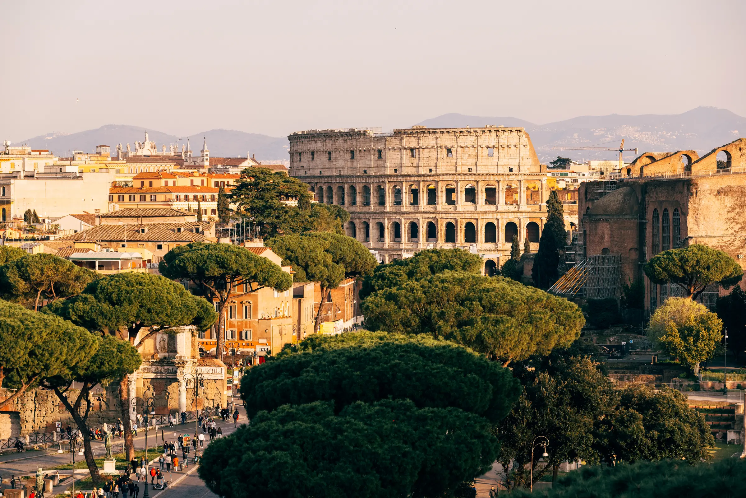 The Colosseum in Rome stands proudly under a clear blue sky, encircled by bustling people, roads, and urban life. Trees and distant mountains frame this iconic symbol amidst modern vibrance.