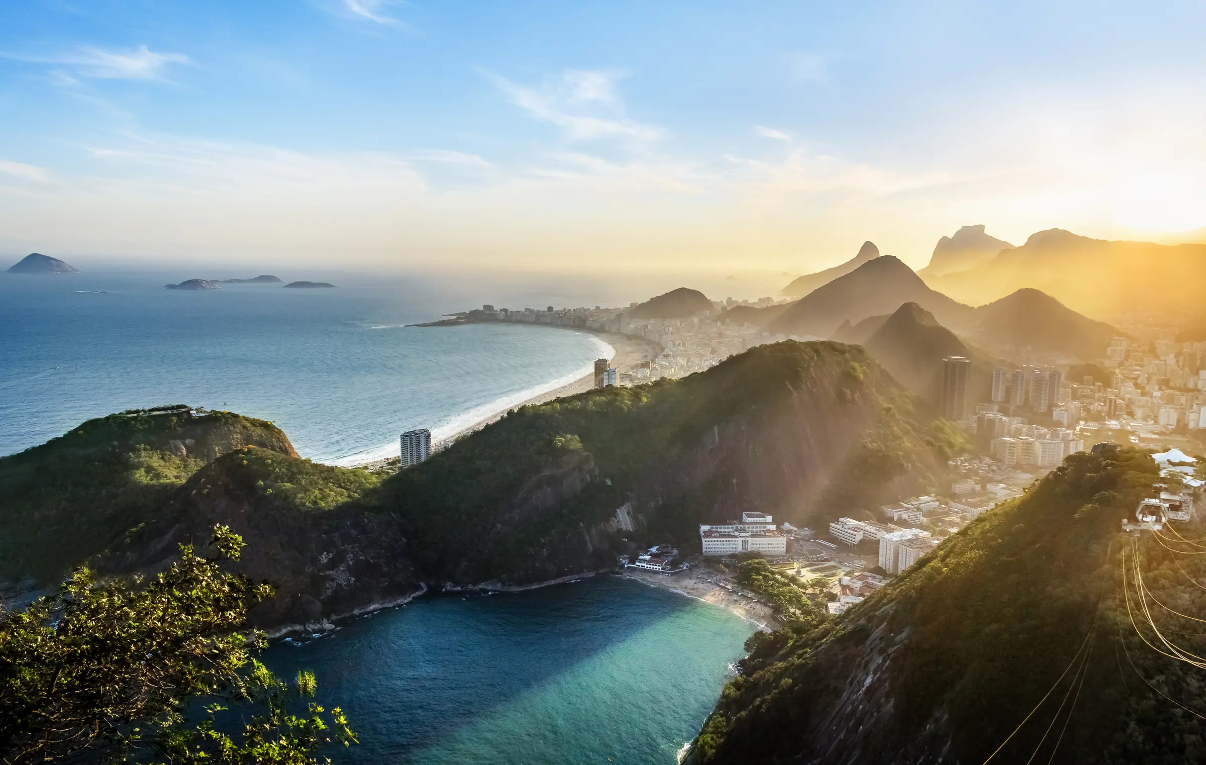 Overlooking Rio from Sugarloaf Mountain, the view is breathtaking: lush hills, city buildings, and the ocean's blue waters. Fluffy clouds dot the sky, enhancing this stunning Brazilian landscape.