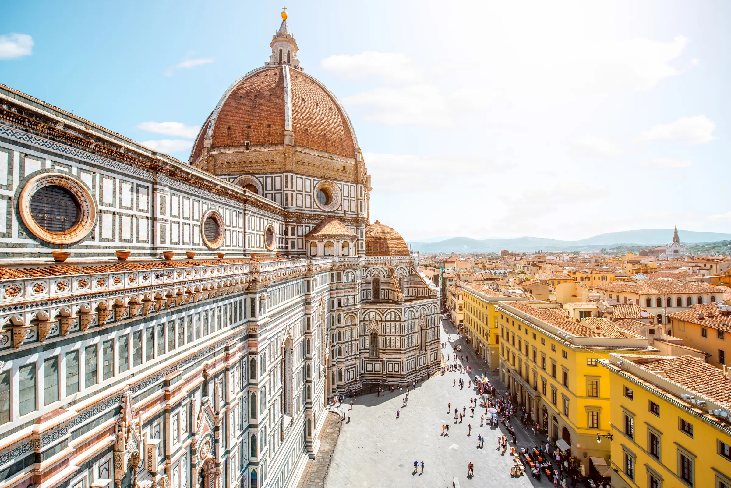 Florence's Cathedral of Santa Maria del Fiore, with its iconic dome, stands against a backdrop of scenic hills and blue skies. Crowds wander below, making it a top romantic getaway spot.
