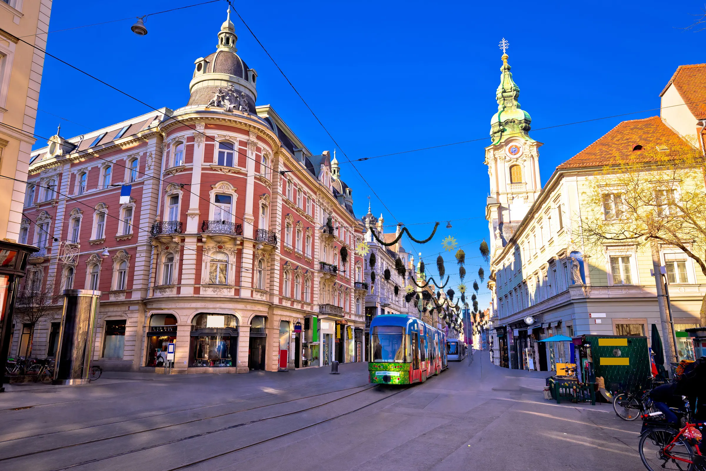 In Graz, a tram glides through a city street, flanked by buildings with windows and trees. A clock tower stands proudly in the backdrop, with bicycles lining the road and the sky peeking above.