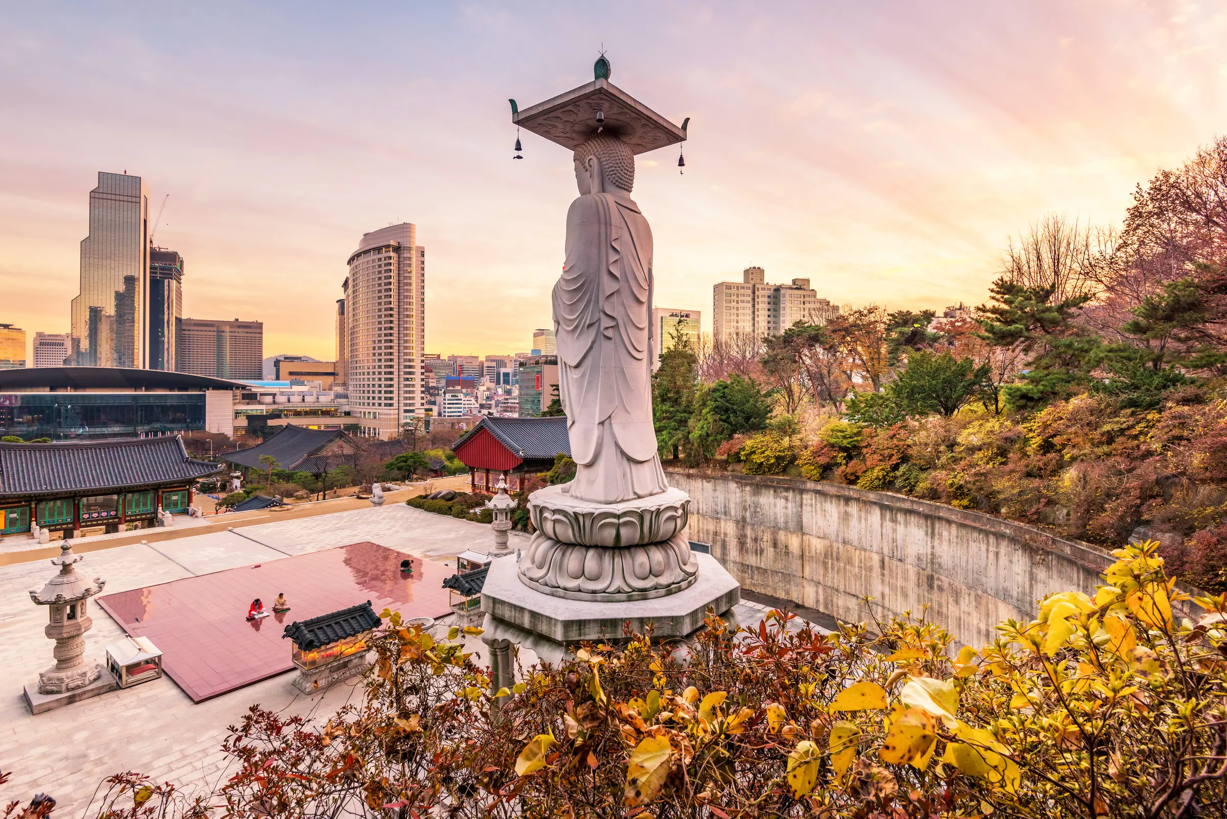 A statue of a woman stands atop a hill in Seoul, surrounded by trees, buildings, sheds, and people on benches. The cloud-filled sky enhances the peaceful ambiance of the scene.