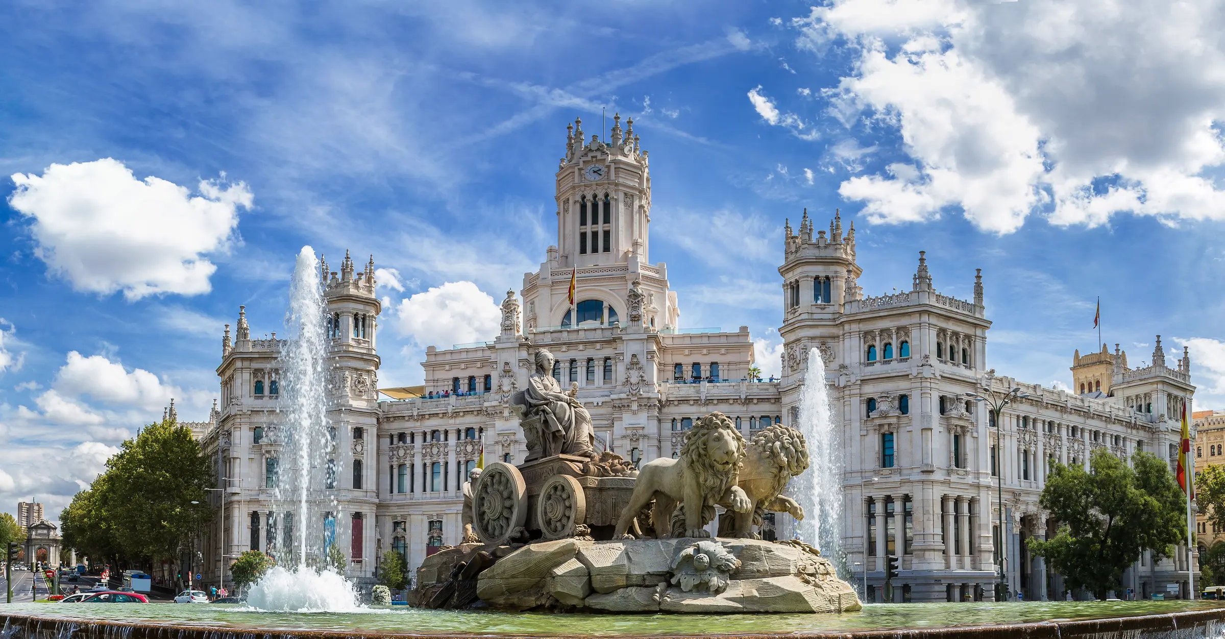 In Madrid, a grand scene unfolds: a clock tower stands tall behind a fountain, surrounded by statues and trees. Vehicles and people fill the bustling streets under a cloudy sky, capturing urban life vividly.
