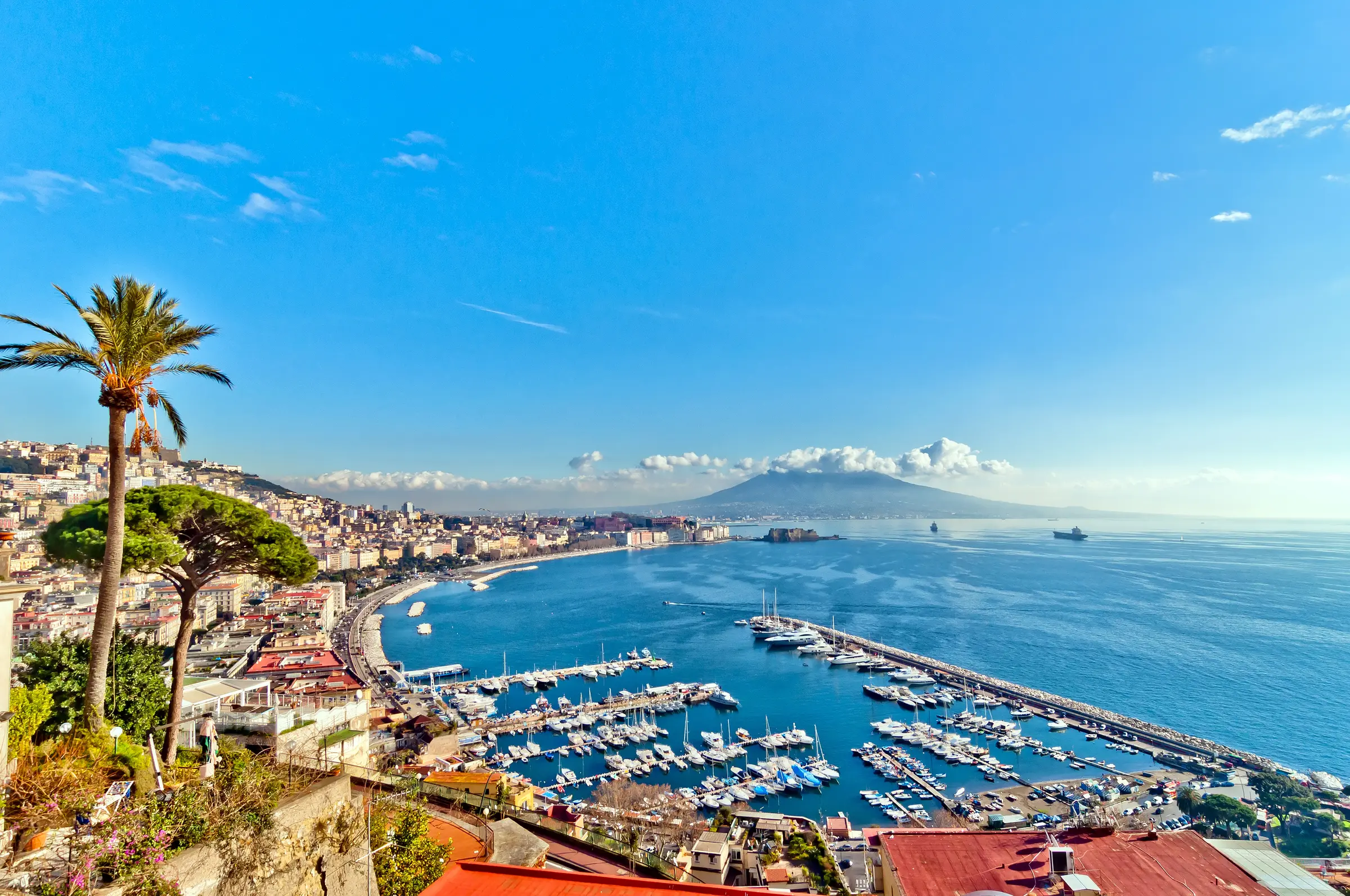 Naples' harbor, vibrant with docked boats, is framed by lush greenery and colorful flowers. Surrounding buildings add charm, while a hill and cloudy sky create a picturesque backdrop.