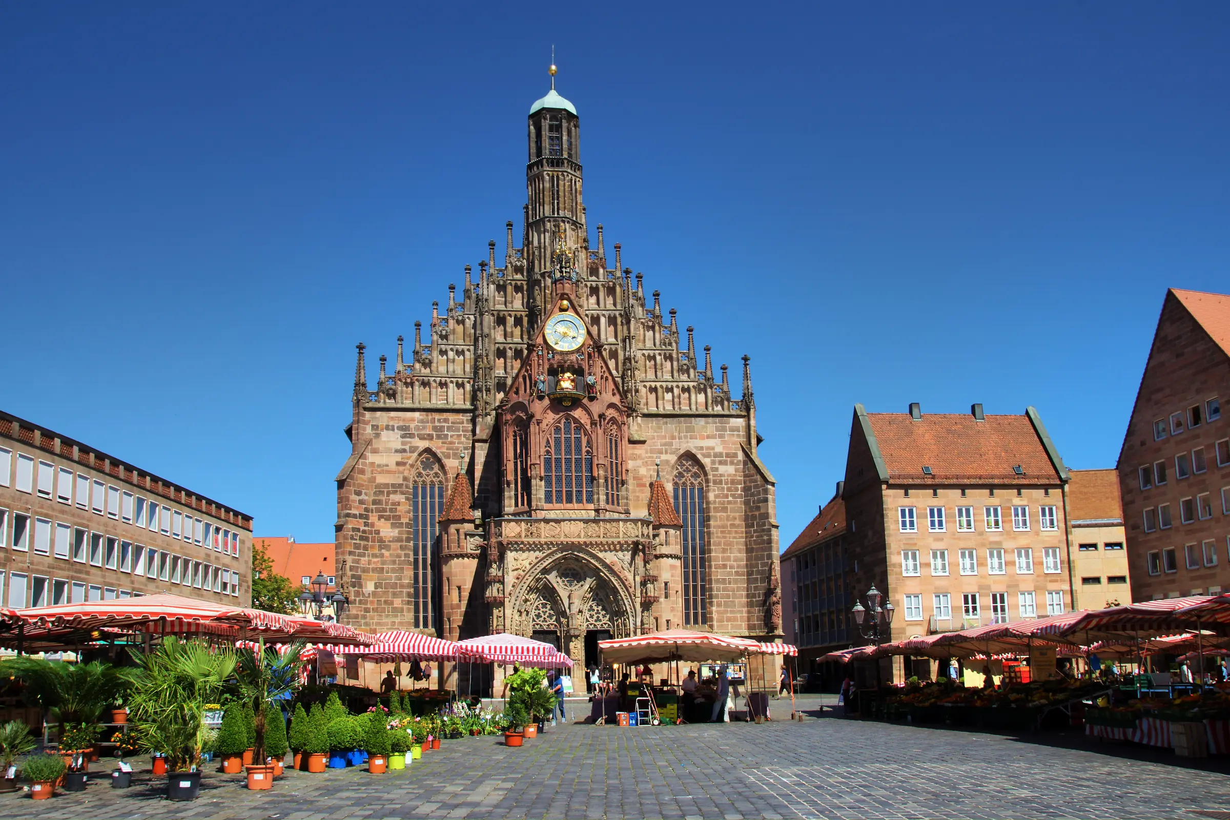 In Nuremberg's lively market square, people gather amidst buildings adorned with windows. A clock tower graces the backdrop, while umbrellas and potted plants dot the scene under a visible sky.