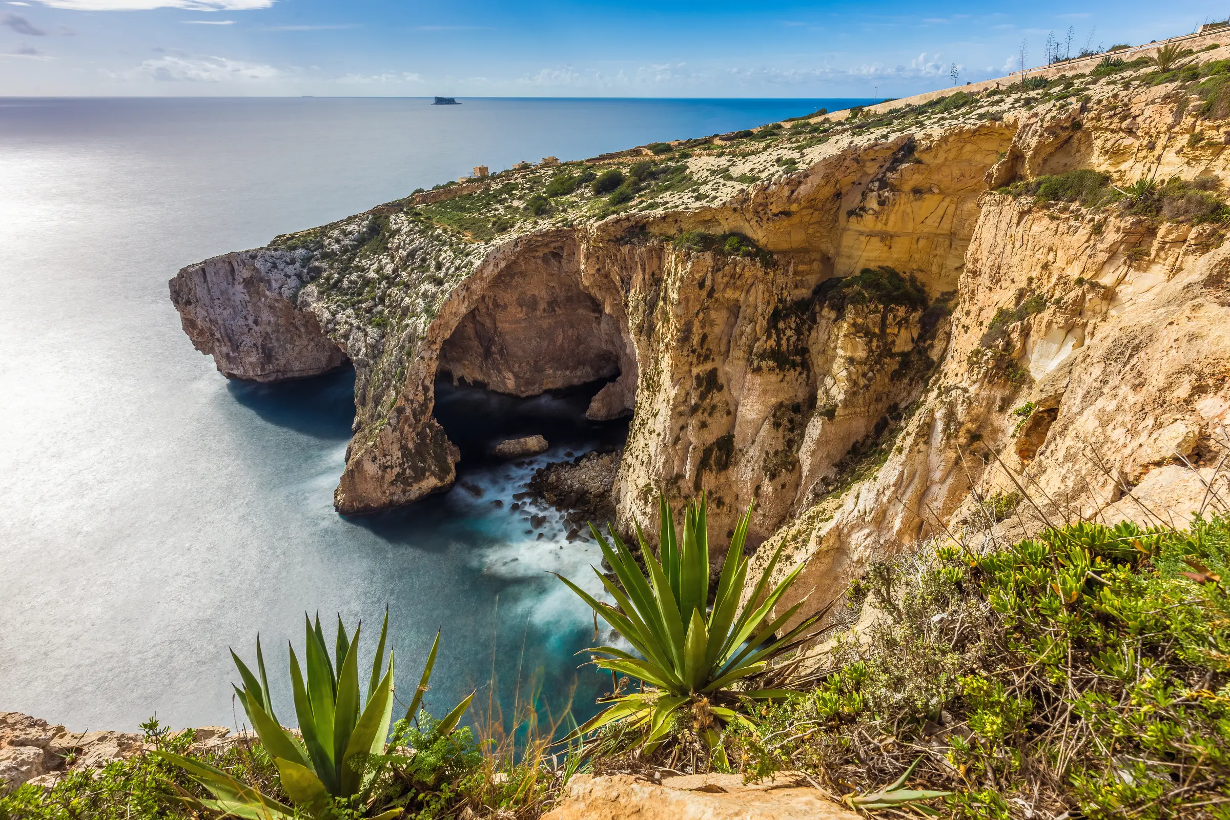 In Luqa, Malta, an image captures Gozo's Azure Window, flanked by rocky cliffs, vibrant plants, and clear water, under a sky adorned with fluffy, white clouds, creating a breathtaking scene.