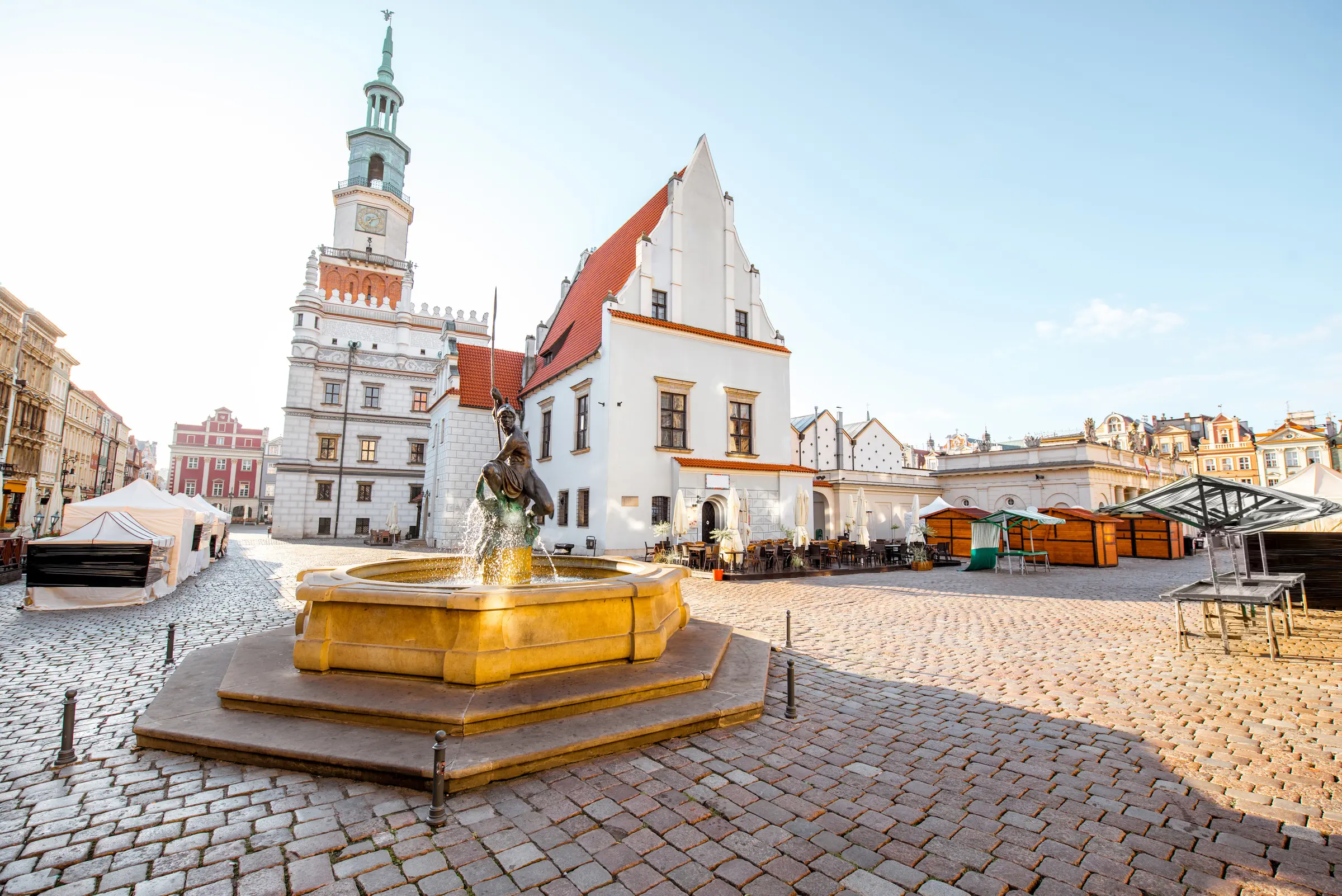 In Poznań, a fountain graces a city square, overlooked by a towering clock. Surrounding buildings host tables and chairs beneath cloud-filled skies, crafting a serene and inviting scene.