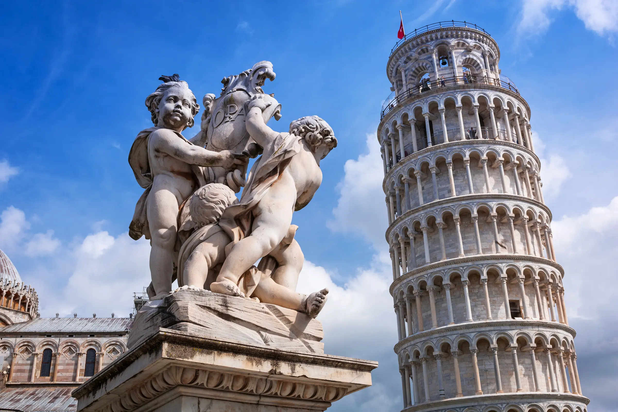 In Pisa, Italy, the iconic Leaning Tower stands with a statue in the foreground. A building completes the scene, set against a backdrop of fluffy white clouds filling the sky—a truly picturesque sight.