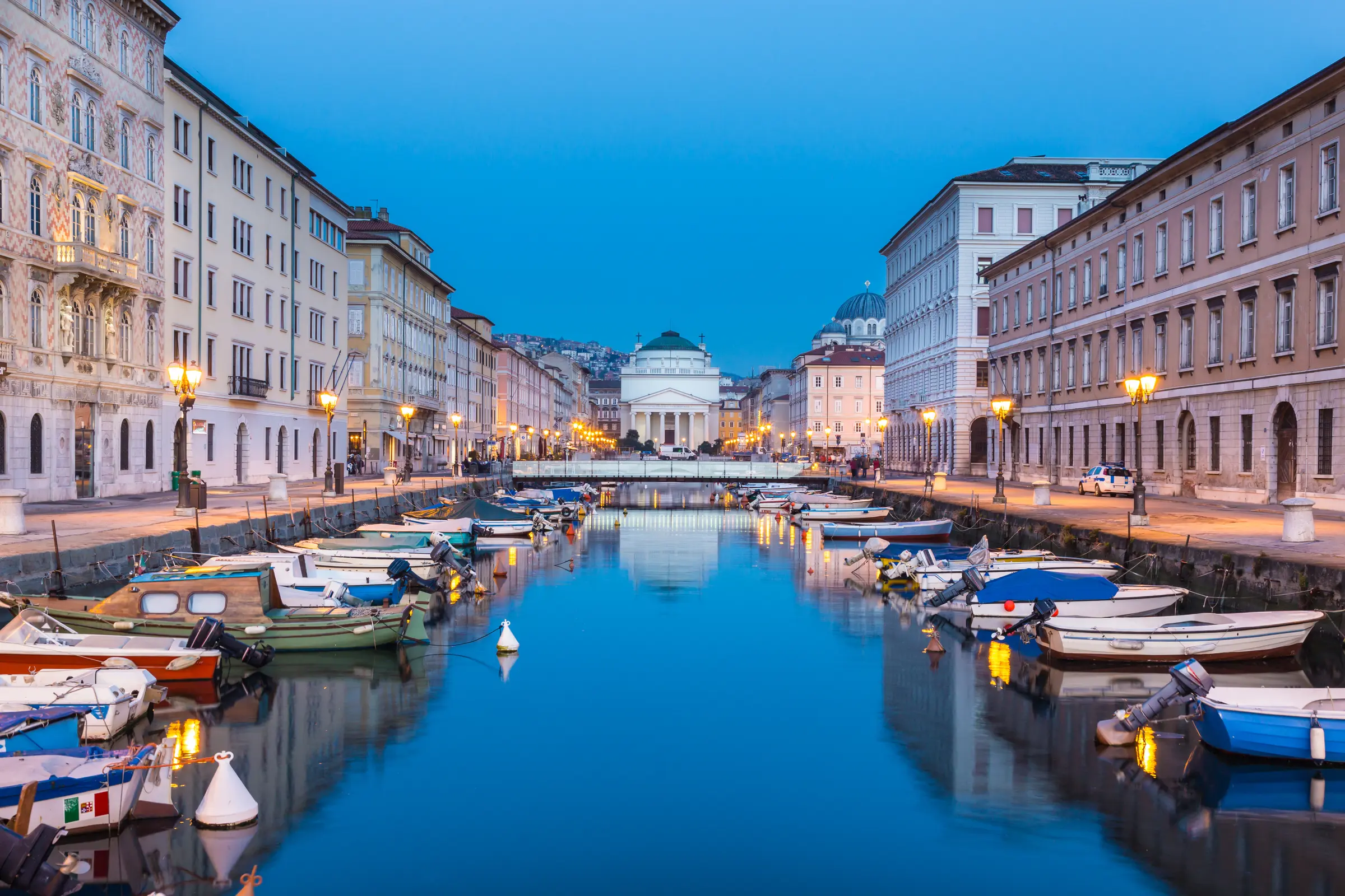 At dusk in Trieste, boats quietly dock by the illuminated city. Street lights glow warmly on buildings, with vehicles moving along the roads, all set against a serene sky backdrop.