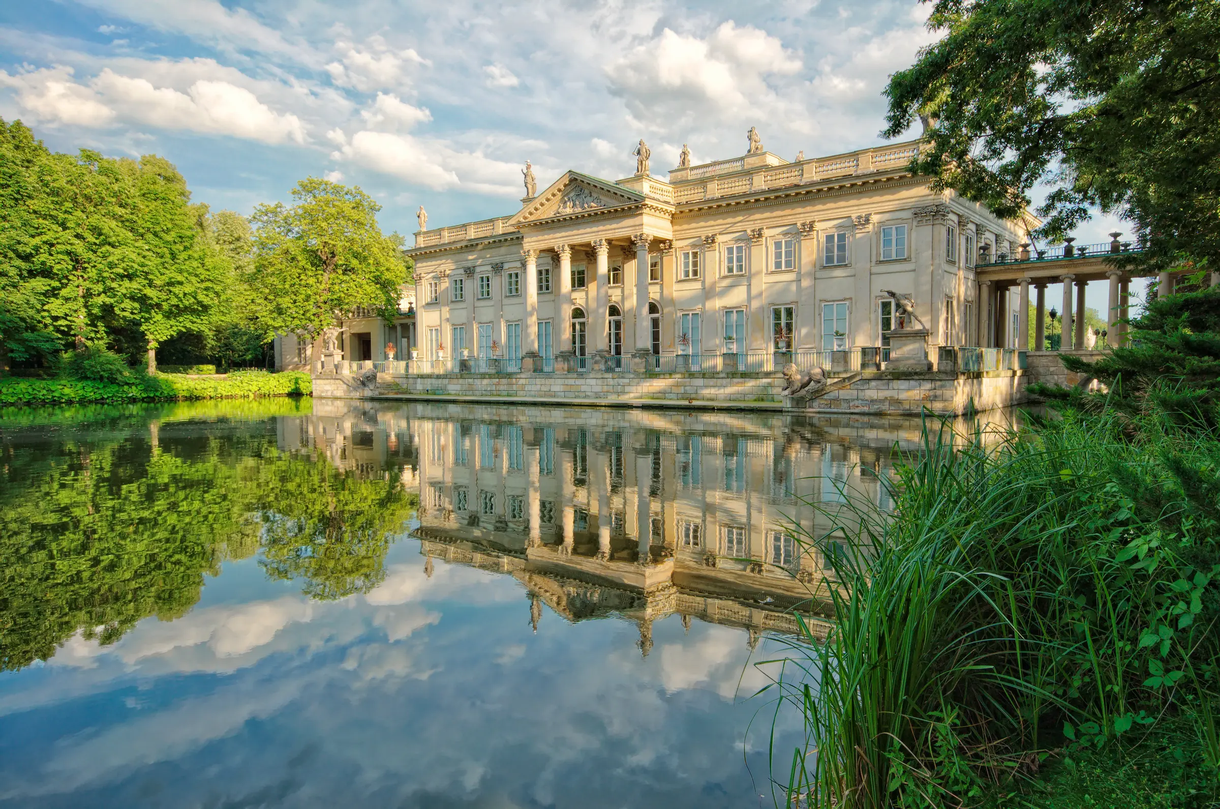 In Warsaw, a grand building with windows, pillars, and statues sits by the water, embraced by lush plants, grass, and a cluster of trees under a cloudy sky, creating a serene, picturesque scene.