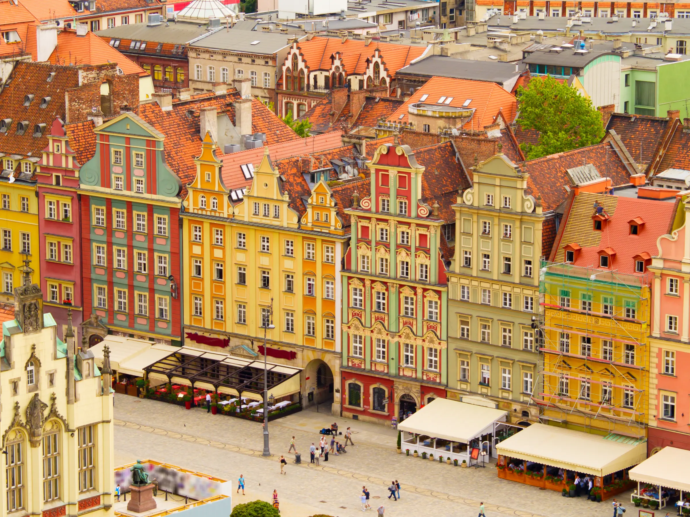 In Wrocław's old town square, discover charming buildings, lush trees, iconic light poles, and a lively crowd. A striking statue on a pedestal adds historical charm to this vibrant scene.