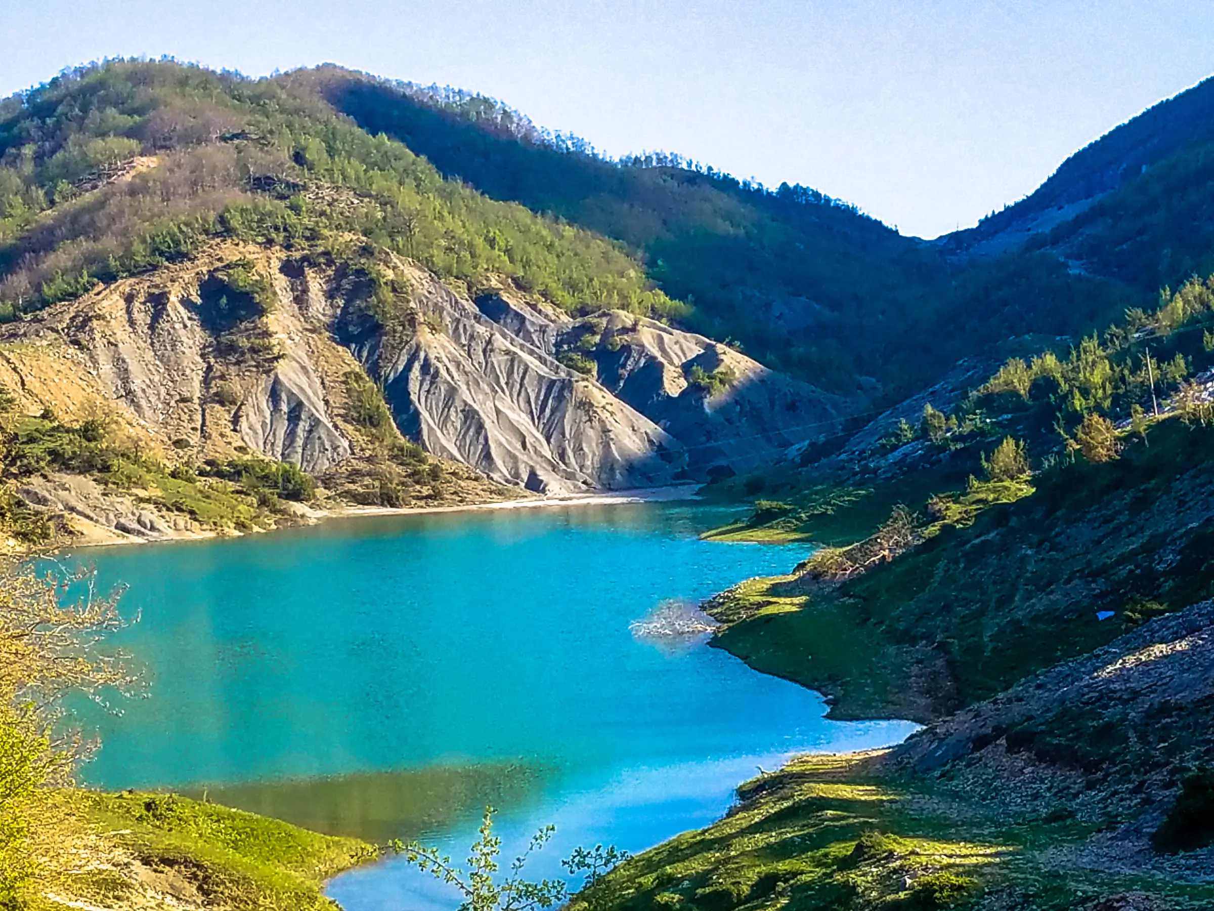 In Tirana, a vast lake is nestled among lush green hills and trees, beneath a bright blue sky, creating a picturesque and serene natural landscape.