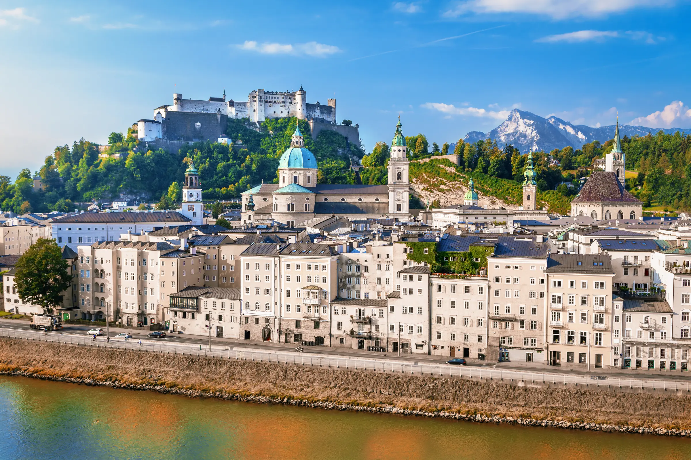 Salzburg, Austria, showcases its charm with buildings amidst lush trees and roads dotted with vehicles. Majestic mountains rise in the background, while a serene body of water reflects the cloudy sky above.