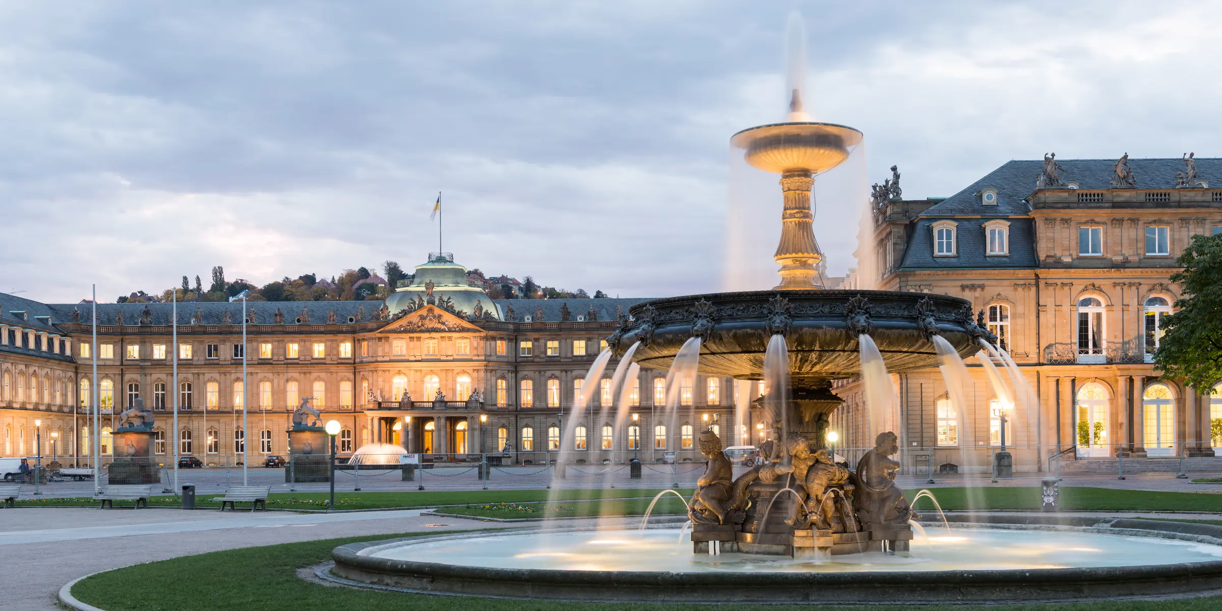 In Stuttgart, a grand building stands adorned with windows and a flag. A fountain graces its front, flanked by statues, light poles, benches, and trees, all beneath a cloudy sky.