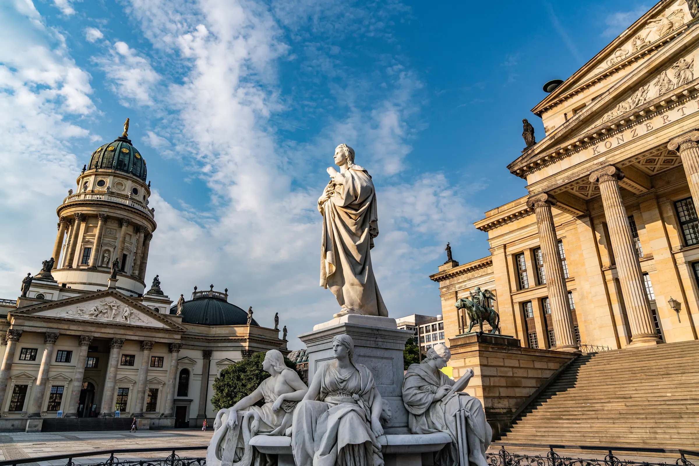 In Berlin, a statue of a woman graces a fountain before an ornate building with pillars, arches, and a dome. Steps and a railing lead up, framed by trees under a cloudy sky.