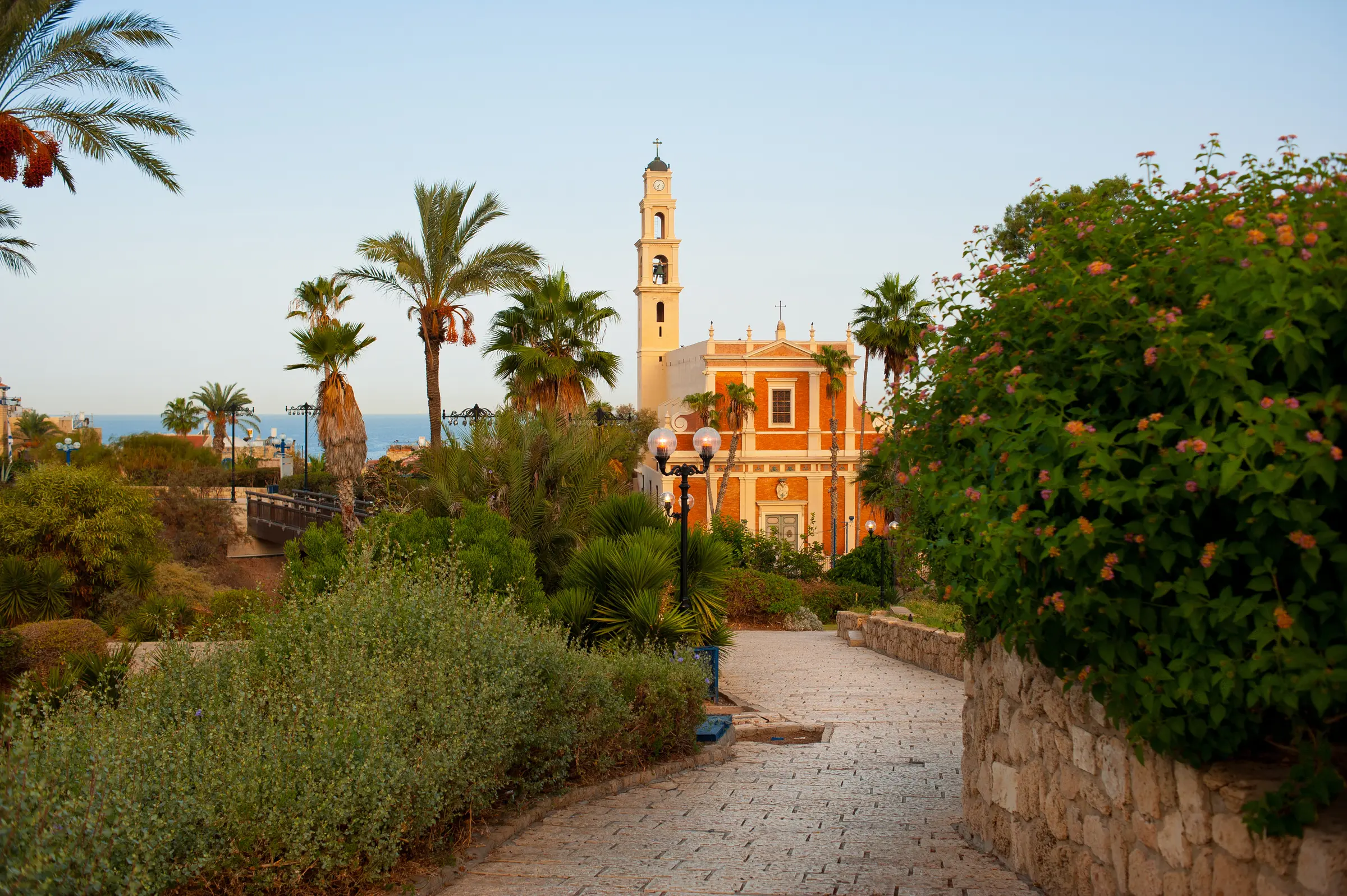In Tel Aviv, a walkway leads to a church with a clock tower, framed by streetlights, trees, flowers, and a wall. The sky stretches above, while a distant body of water adds to the scenic view.