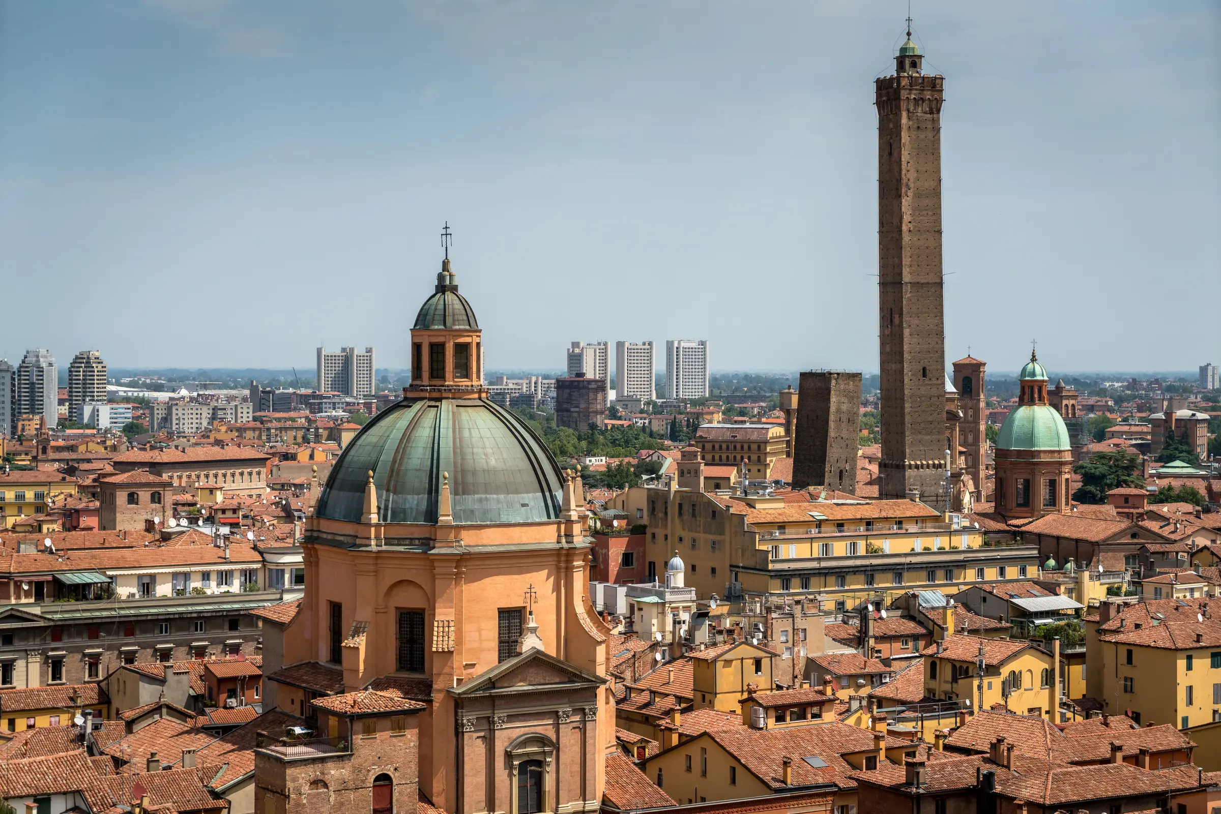 A scenic view from a hilltop reveals Bologna's vibrant cityscape, framed by lush trees and set against a clear blue sky. The scene captures the blend of urban charm and natural beauty.