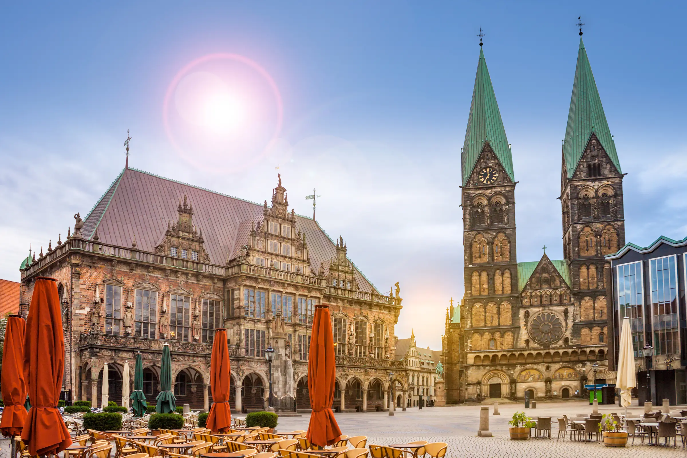 In Bremen, a grand building with a clock tower stands amid tables, chairs, and plants. The sunny sky, dotted with clouds, enhances the lively atmosphere, captured perfectly in this picturesque setting.