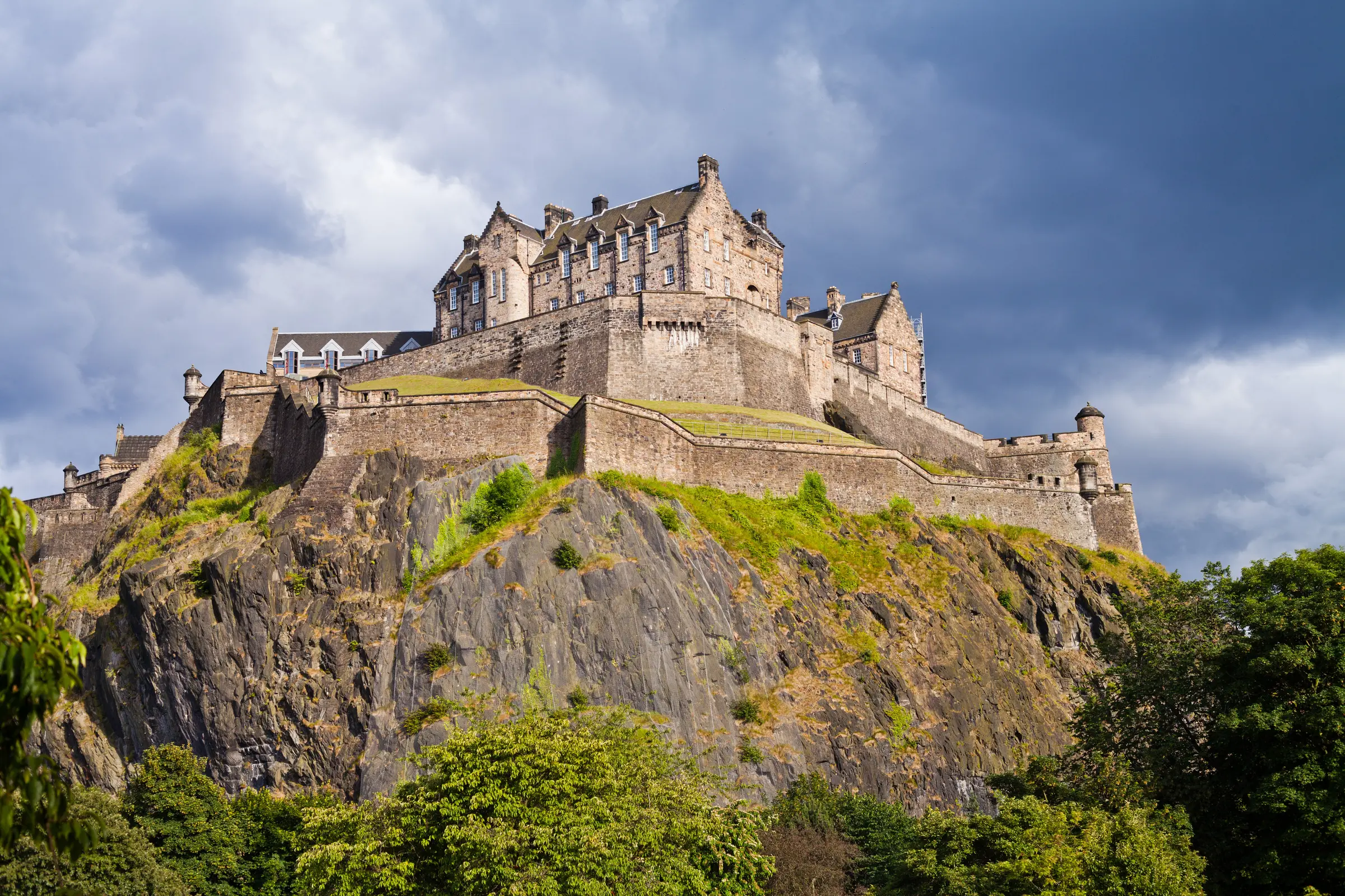 Edinburgh Castle stands majestically on a hill in Scotland, enveloped by verdant grass and trees. A dramatic cloudy sky enhances its timeless allure, creating a striking silhouette in Edinburgh's landscape.