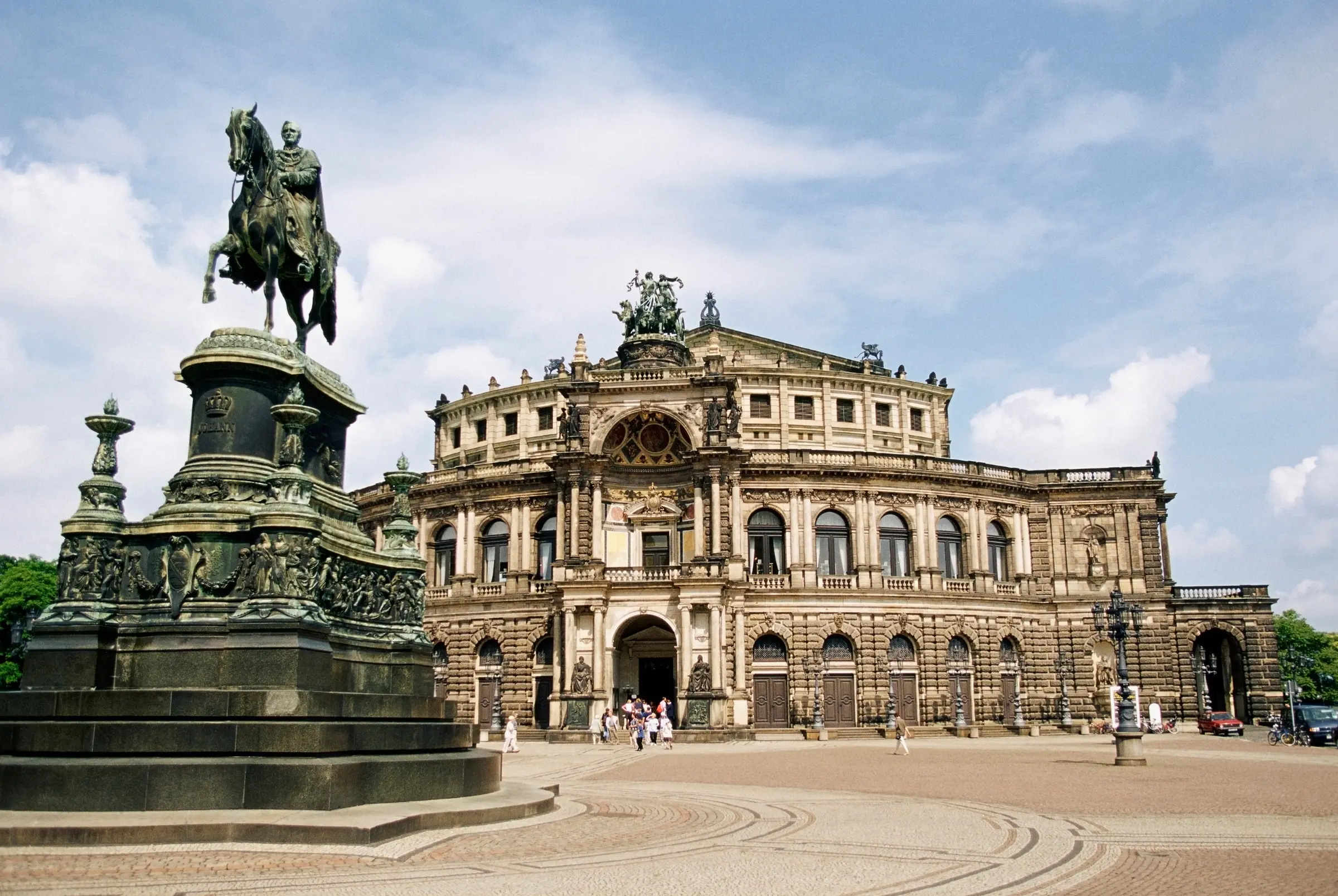 In Dresden, a grand building stands alongside a statue of a man on horseback. The scene is vibrant, with streetlight poles, vehicles, people, and trees under a cloudy sky—all bustling with life.