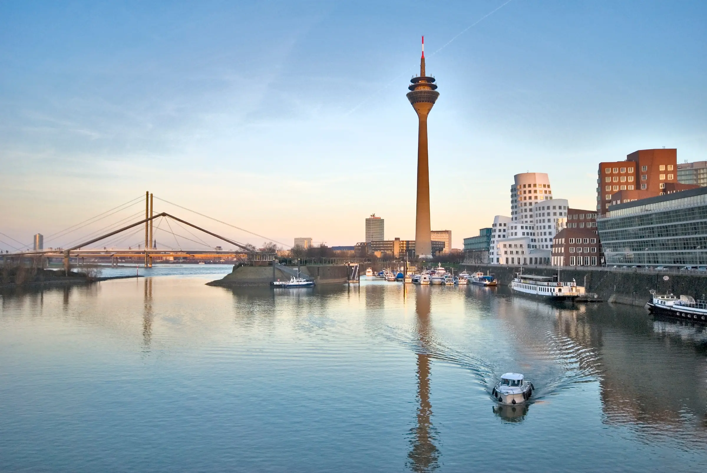 Düsseldorf features a scenic river view with boats and a bridge adorned with poles and ropes. Trees line the banks, flanked by buildings with windows, while a distant tower rises under a cloudy sky.