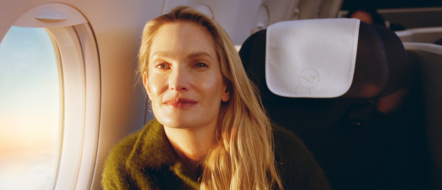 Eine blonde Frau mit langen Haaren sitzt im Flugzeug, sie schaut direkt in die Kamera und ihr Gesicht wird durch das Fenster von der Sonne angestrahlt.