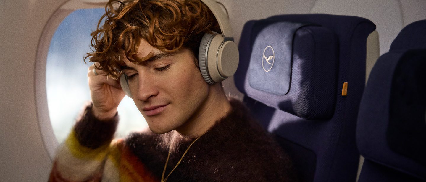 A young man with short red curls and a striped sweater is sitting on the plane wearing headphones.