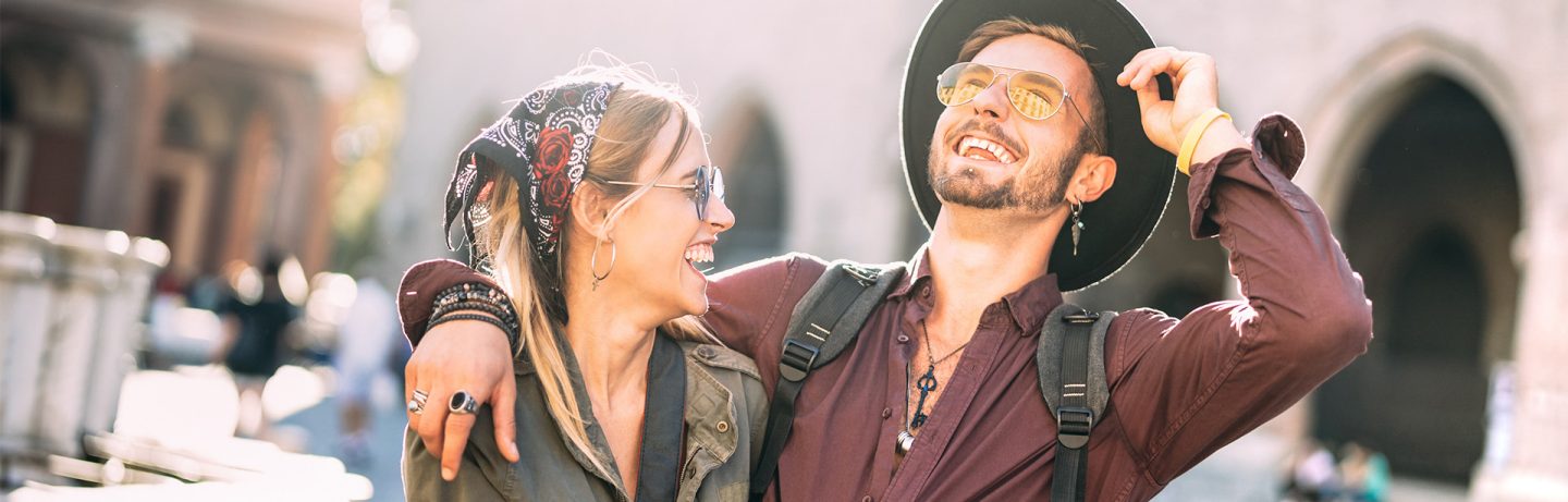 A smiling young man in a hat and sunglasses puts his arm around a young woman wearing sunglasses and a headscarf.  