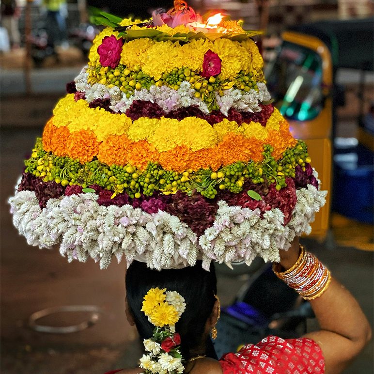 Woman from behind, wearing large colorful flower decoration with candle on her head