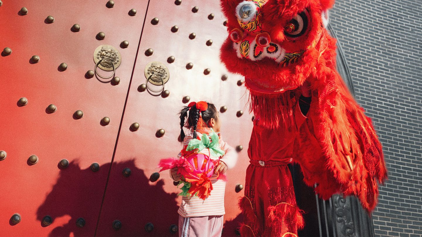 A little Chinese girl with a person dressed as a red dragon in front of a red door. 