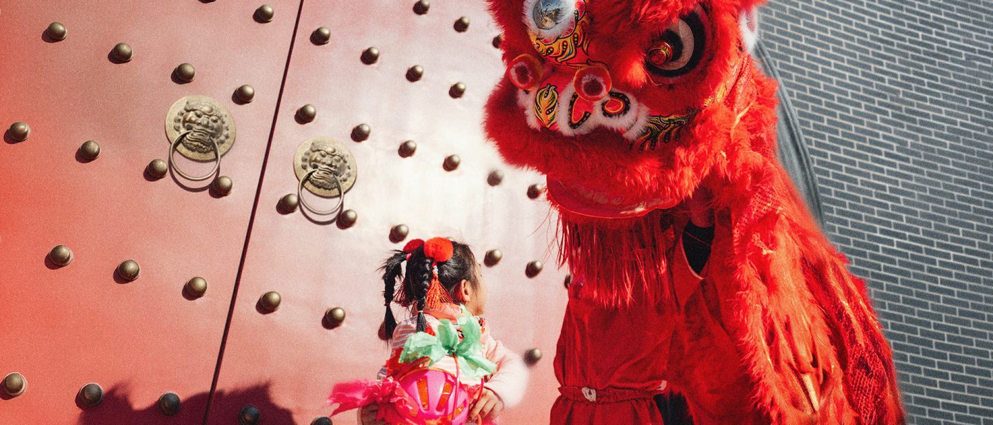 A little Chinese girl with a person dressed as a red dragon in front of a red door. 