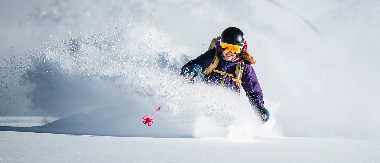 A person skis through deep powder snow, with snow spraying around them. They are wearing a helmet, ski goggles, and a purple ski jacket.