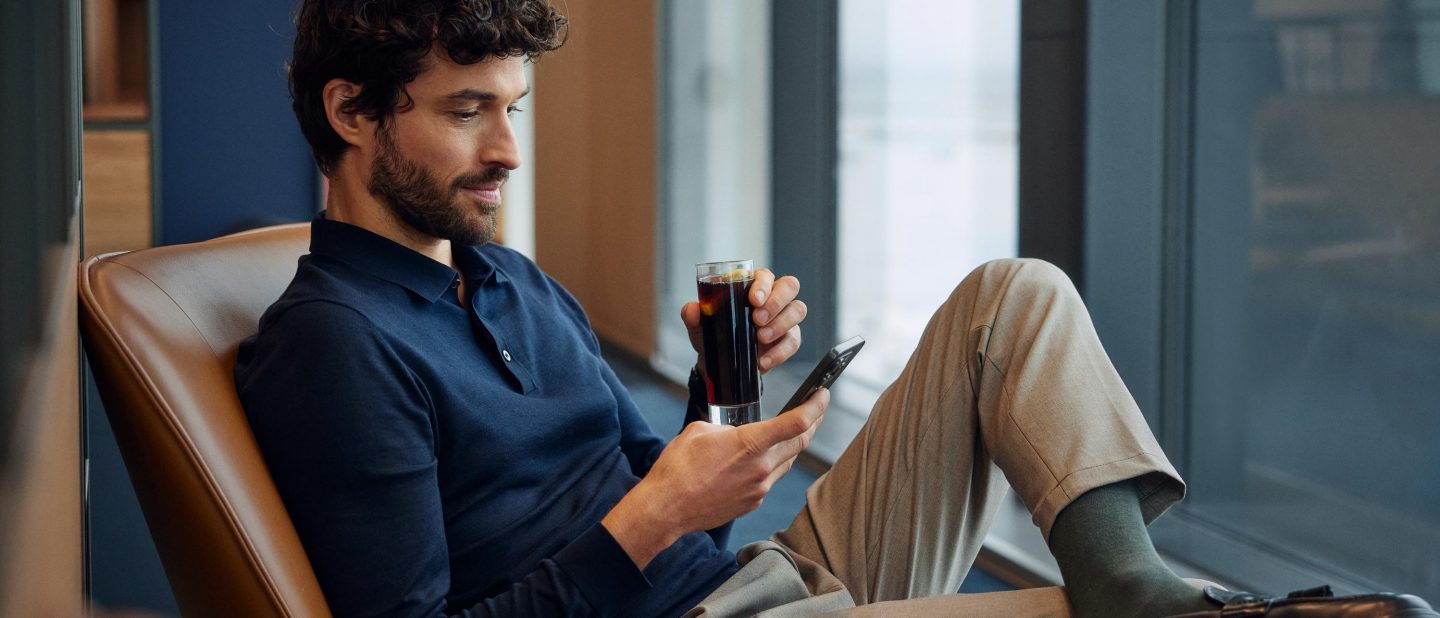 A man sits relaxed on a chair and holds a drink in one hand and a smartphone in the other.