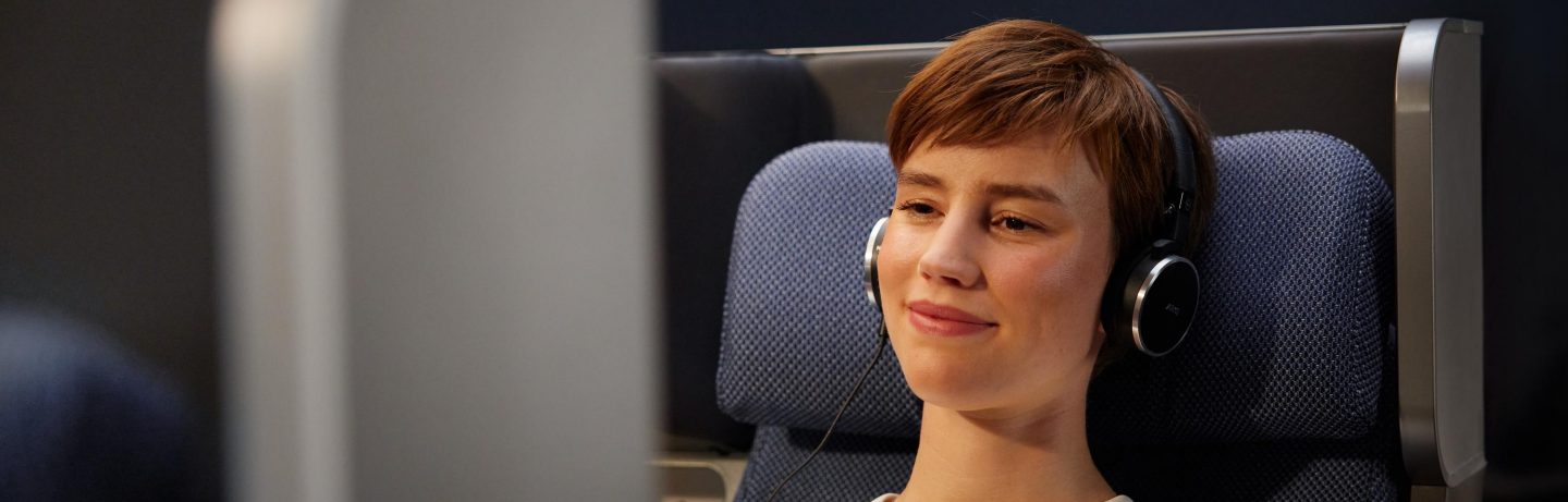 Eine Frau sitzt entspannt in der Allegris Premium Economy Class. Dabei hat sie Kopfh&ouml;rer auf. // A woman sits relaxed in Allegris Premium Economy Class. She has her headphones on.