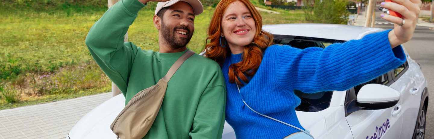 A couple leans against a Free2move car and takes a selfie.